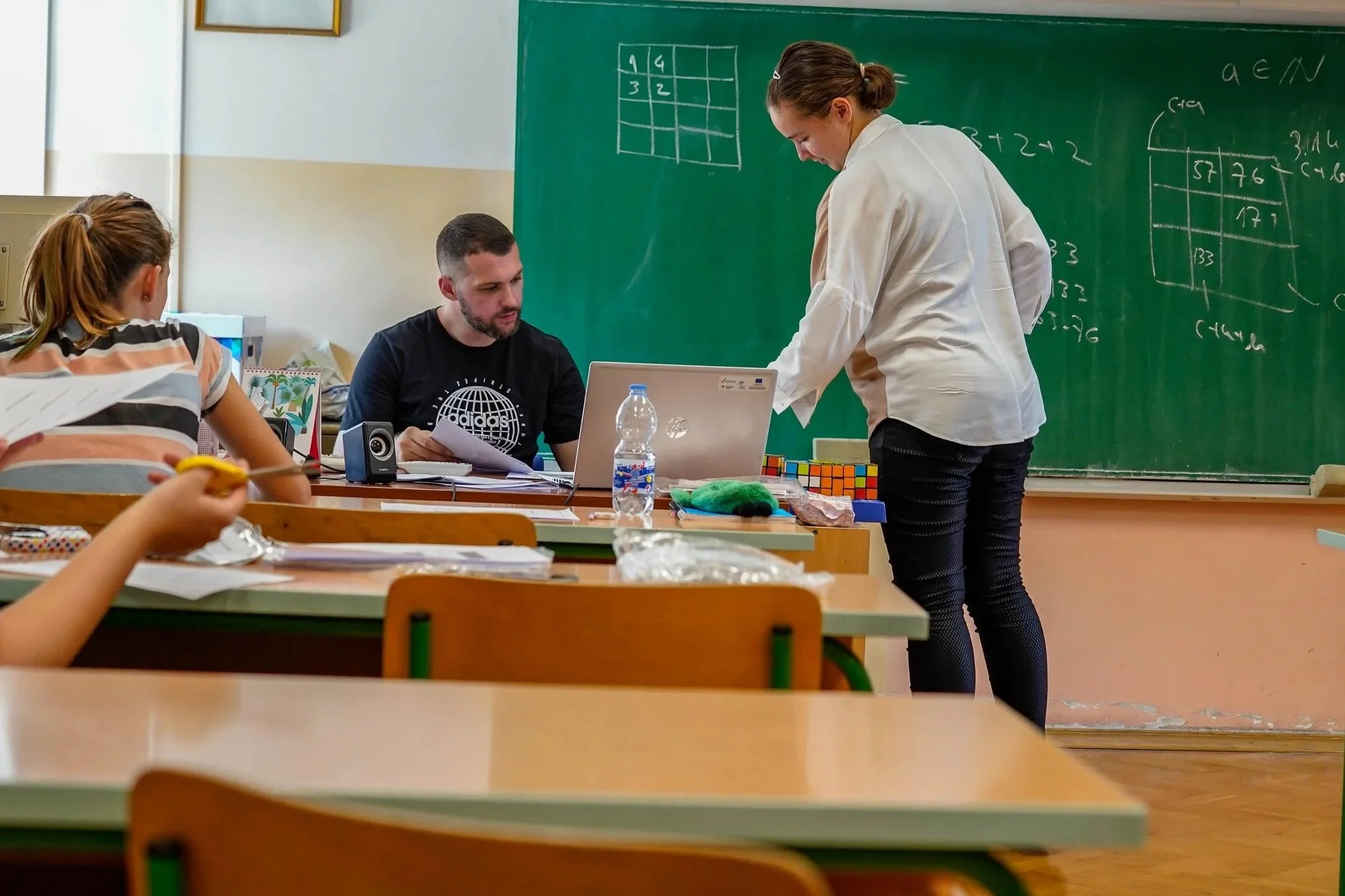 A classroom scene with students and a teacher near a green chalkboard. One female student is standing and explaining something, while two students are seated at desks. The chalkboard has math problems and a tic-tac-toe game drawn on it.