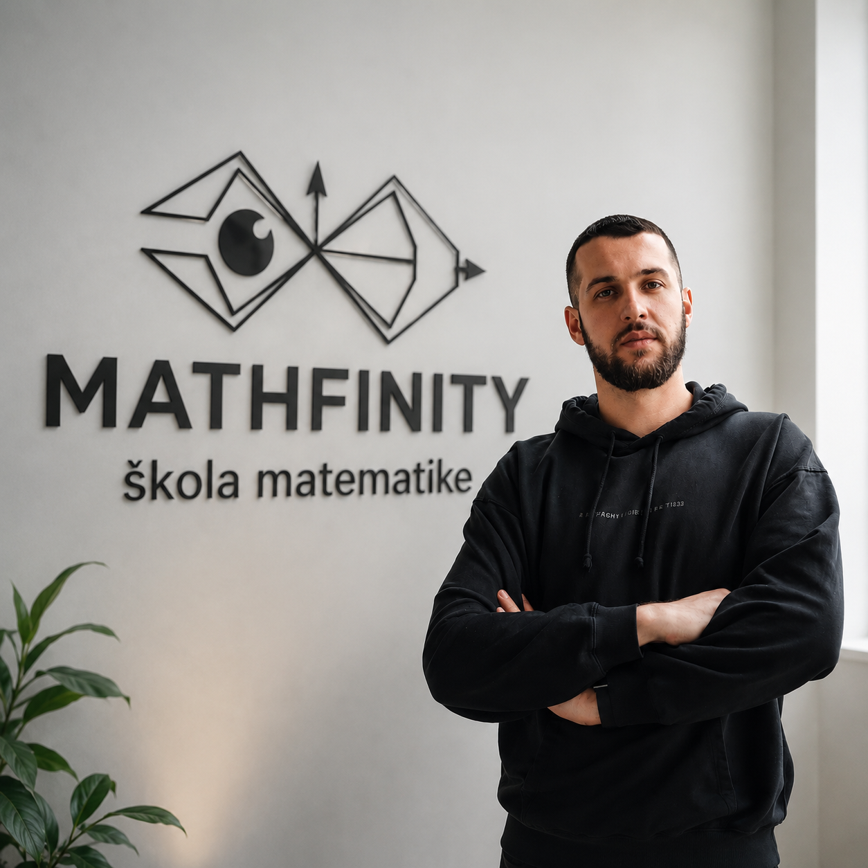 A man with a beard and short hair stands with arms crossed in front of a wall displaying the logo and name of Mathfinity, a math school, with a potted plant in the foreground.