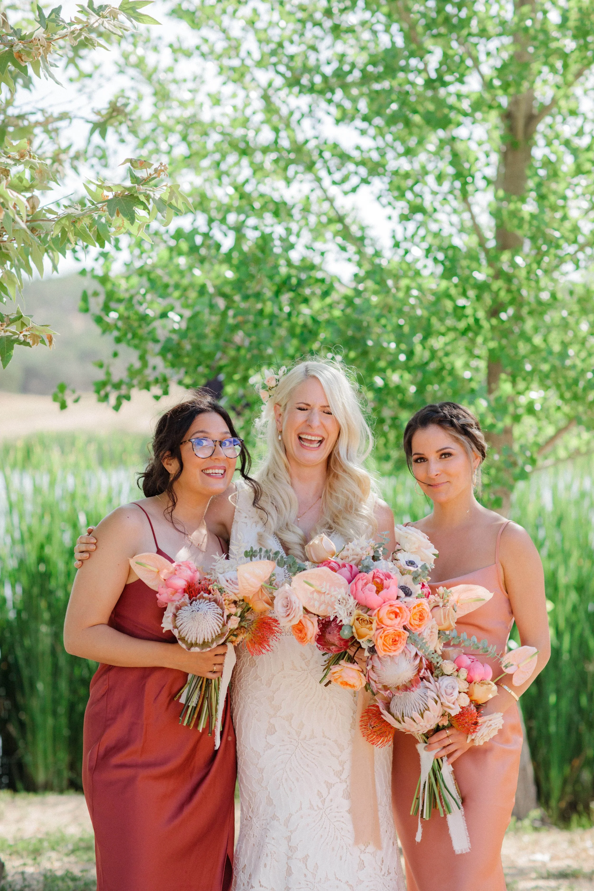 Three women at a wedding, the bride and her bridesmaids, holding bouquets of flowers outdoors under a tree with green leaves.
