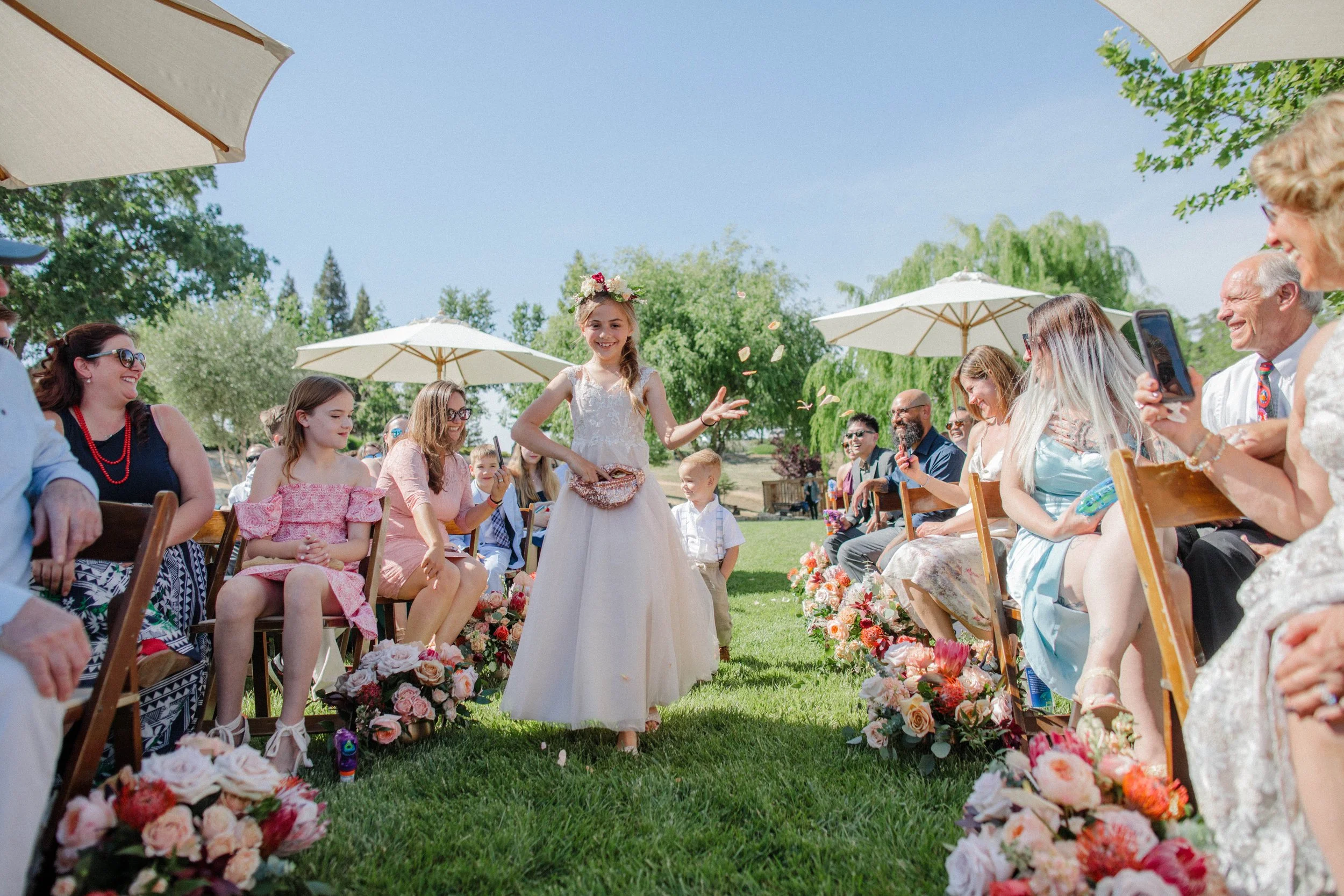 A young girl in a white dress with a flower crown participates in a wedding ceremony outdoors, surrounded by seated guests, under umbrellas on a sunny day.