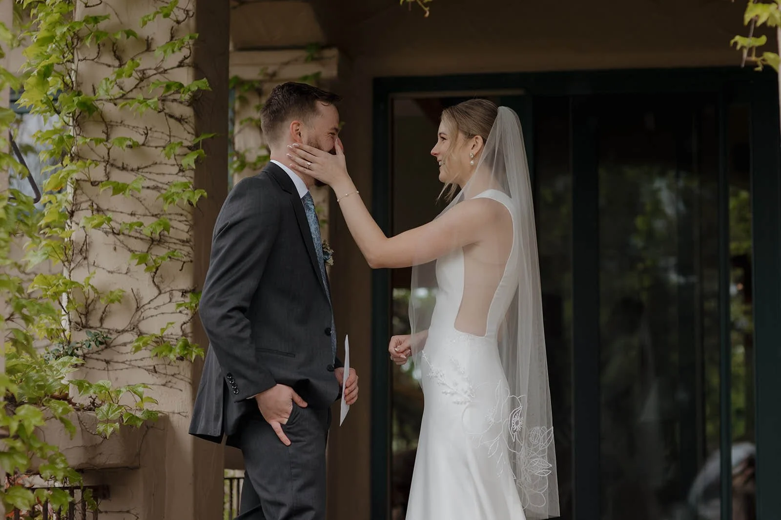A bride and groom are sharing a moment outside a building during their wedding. The bride is touching the groom's face with her hand, and both are smiling.