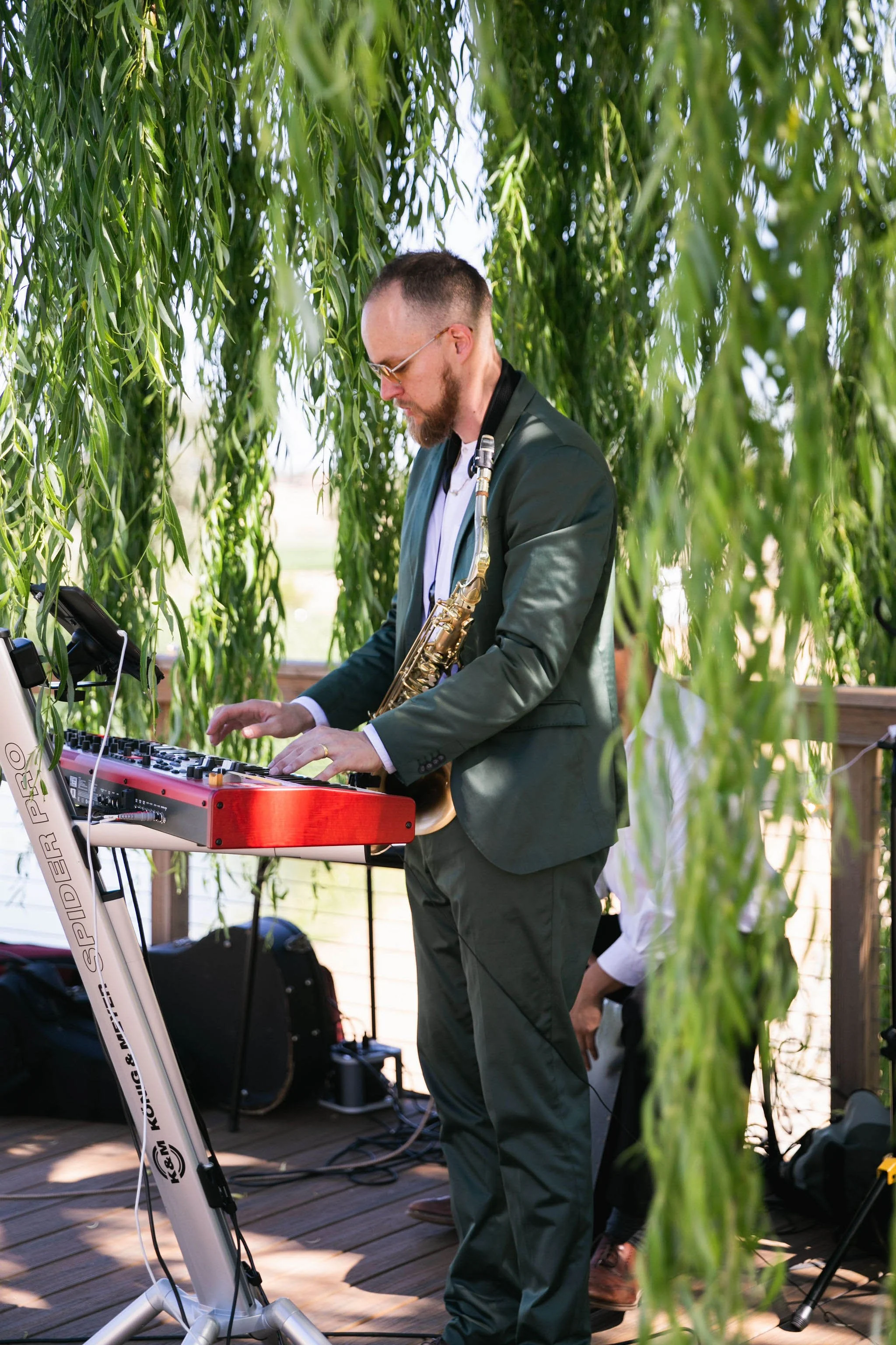 A man in a dark green suit playing a red electronic keyboard and wearing a saxophone around his neck, outdoors under hanging tree branches during daytime.