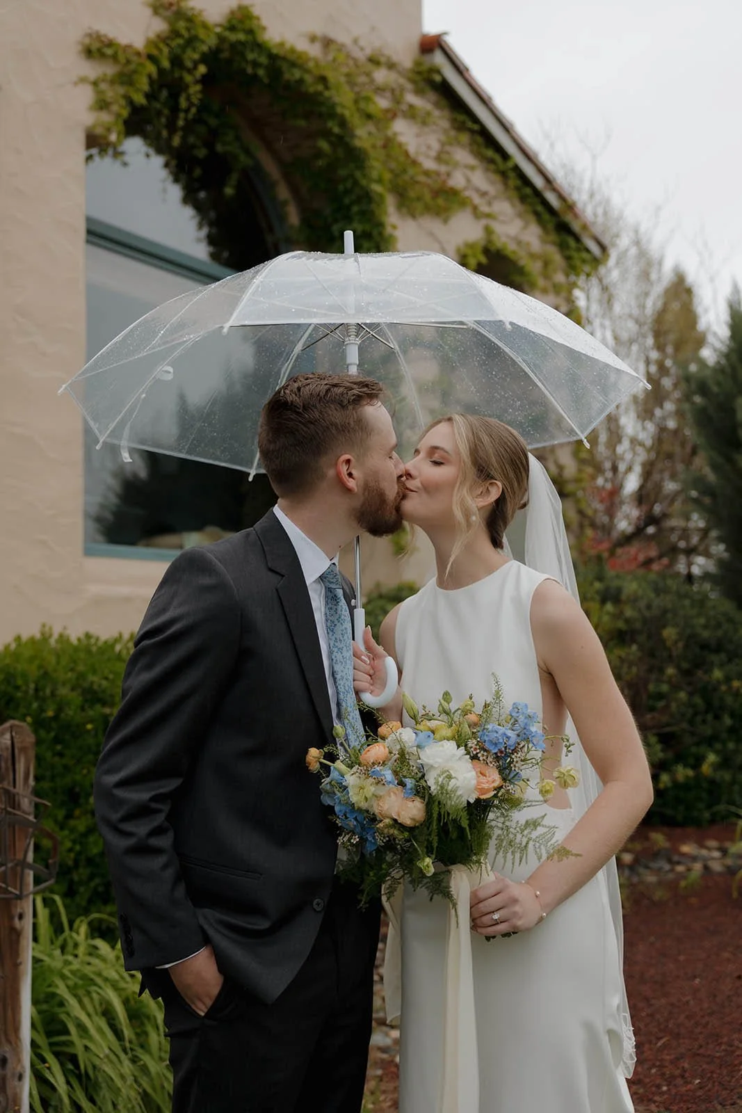 A bride and groom kiss under a clear umbrella outside on a rainy day, with the bride holding a bouquet of flowers.