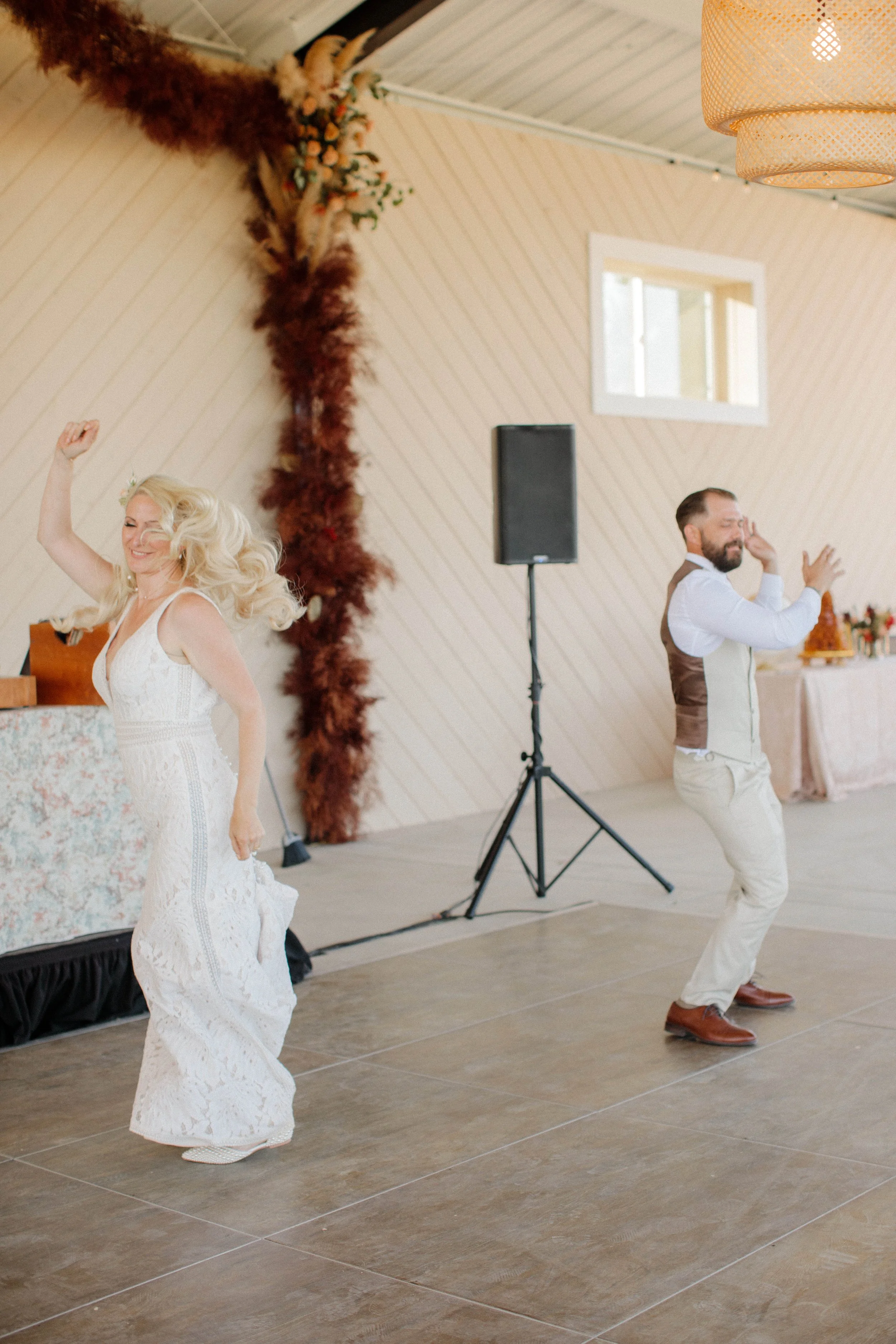 A woman in a white lace dress and a man in a white shirt and brown vest dancing at a wedding reception in a decorated hall.