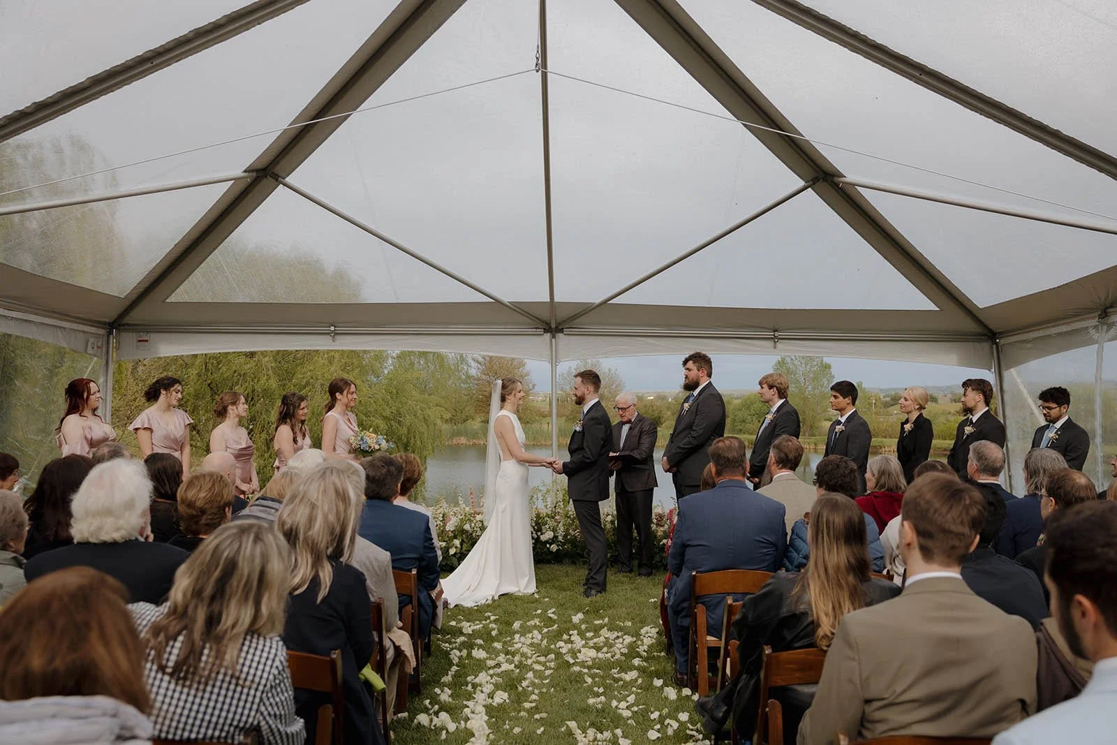 Wedding ceremony under a large outdoor tent by a lake, with the bride and groom holding hands facing each other, surrounded by bridesmaids and groomsmen, and seated guests watching.