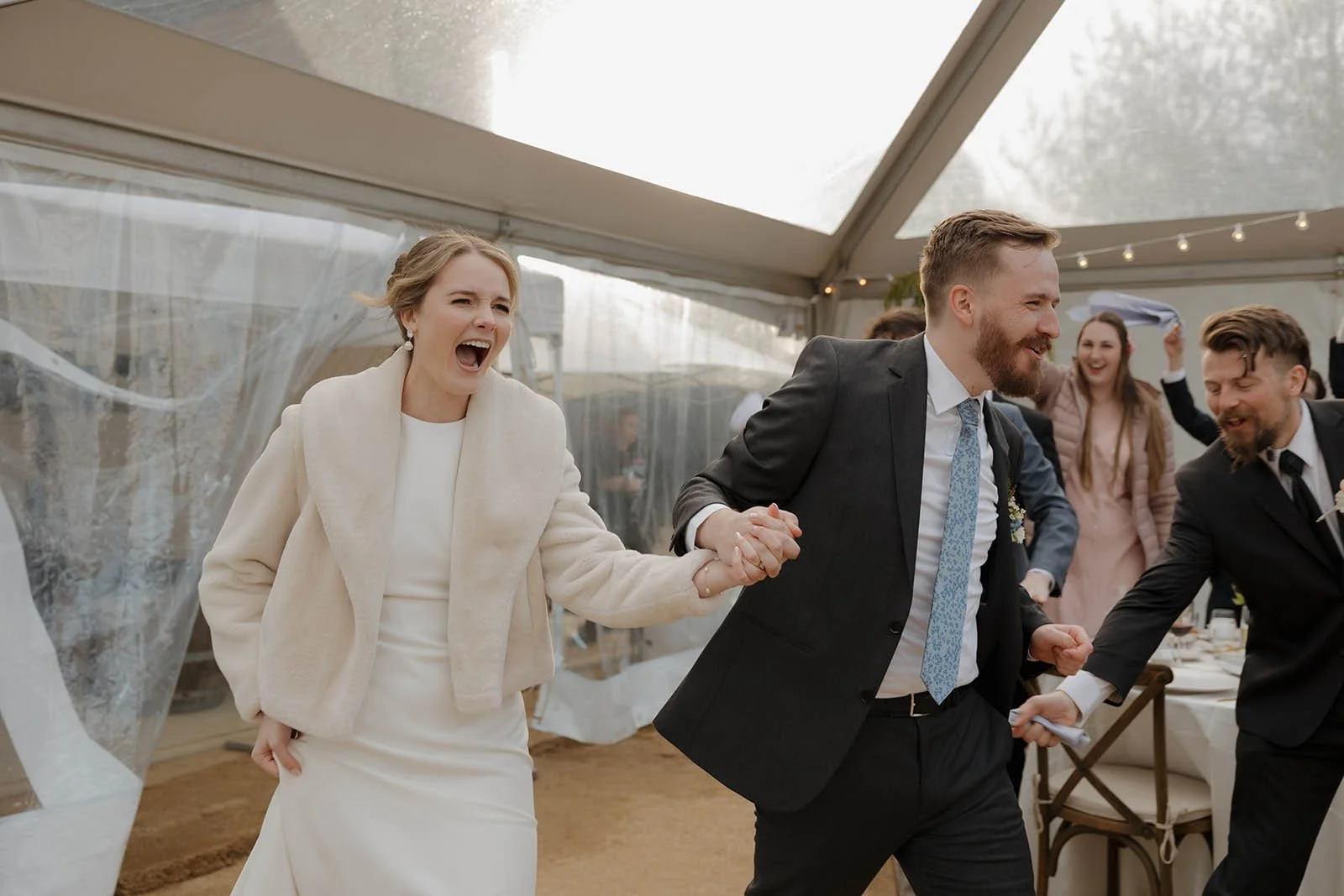 A joyful bride and groom holding hands and dancing at their wedding reception inside a tent, surrounded by friends.