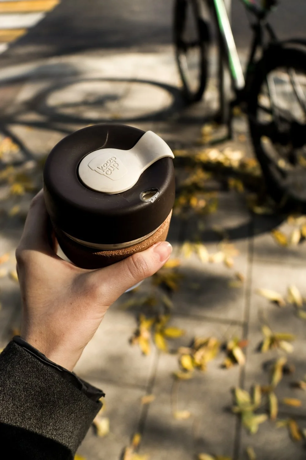 Hand holding a reusable coffee cup with a black lid outdoors, with bicycles and fallen yellow leaves in the background.