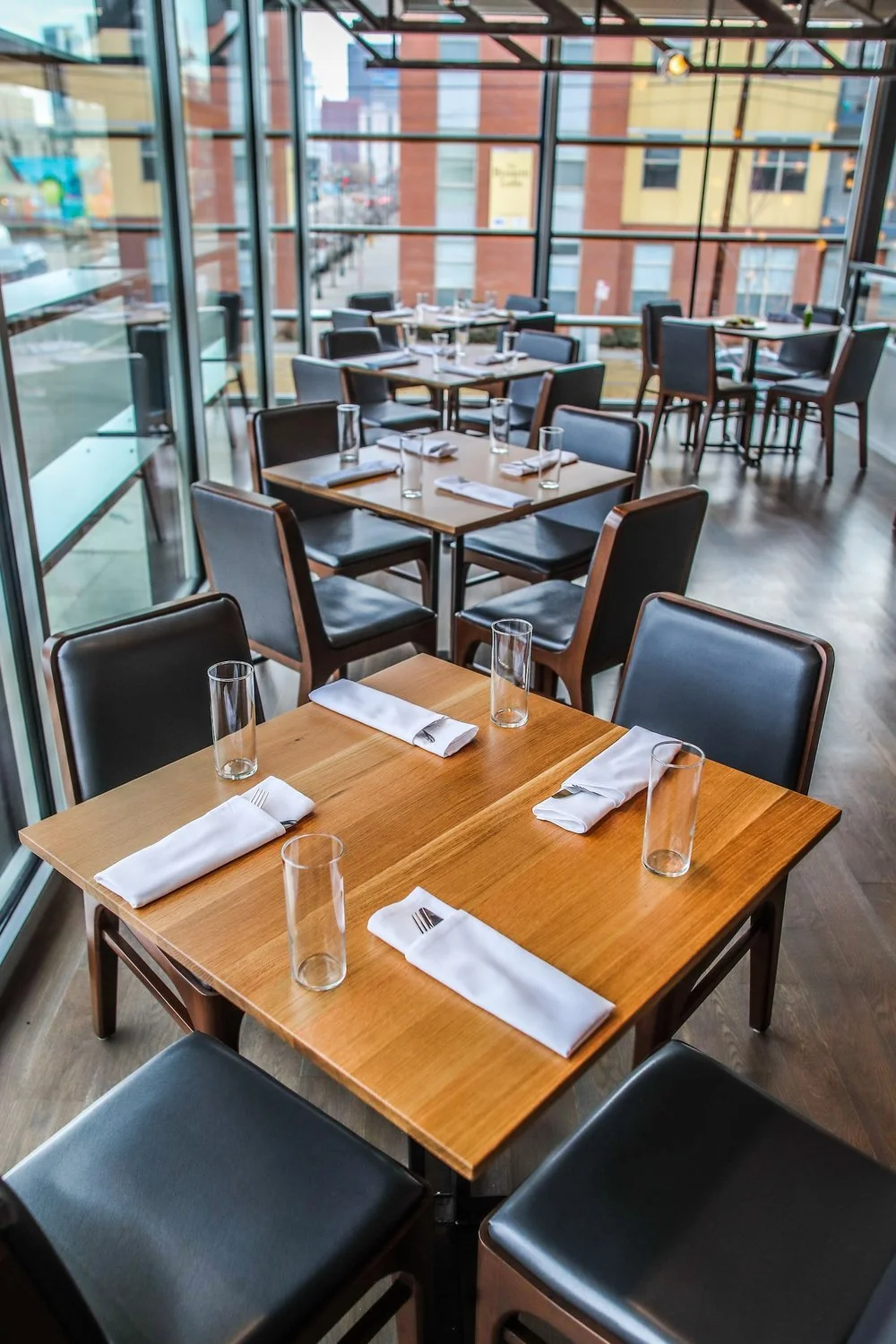 Dining area in a restaurant with wooden tables set with white napkins, forks, and empty glasses, large windows showing city buildings outside.