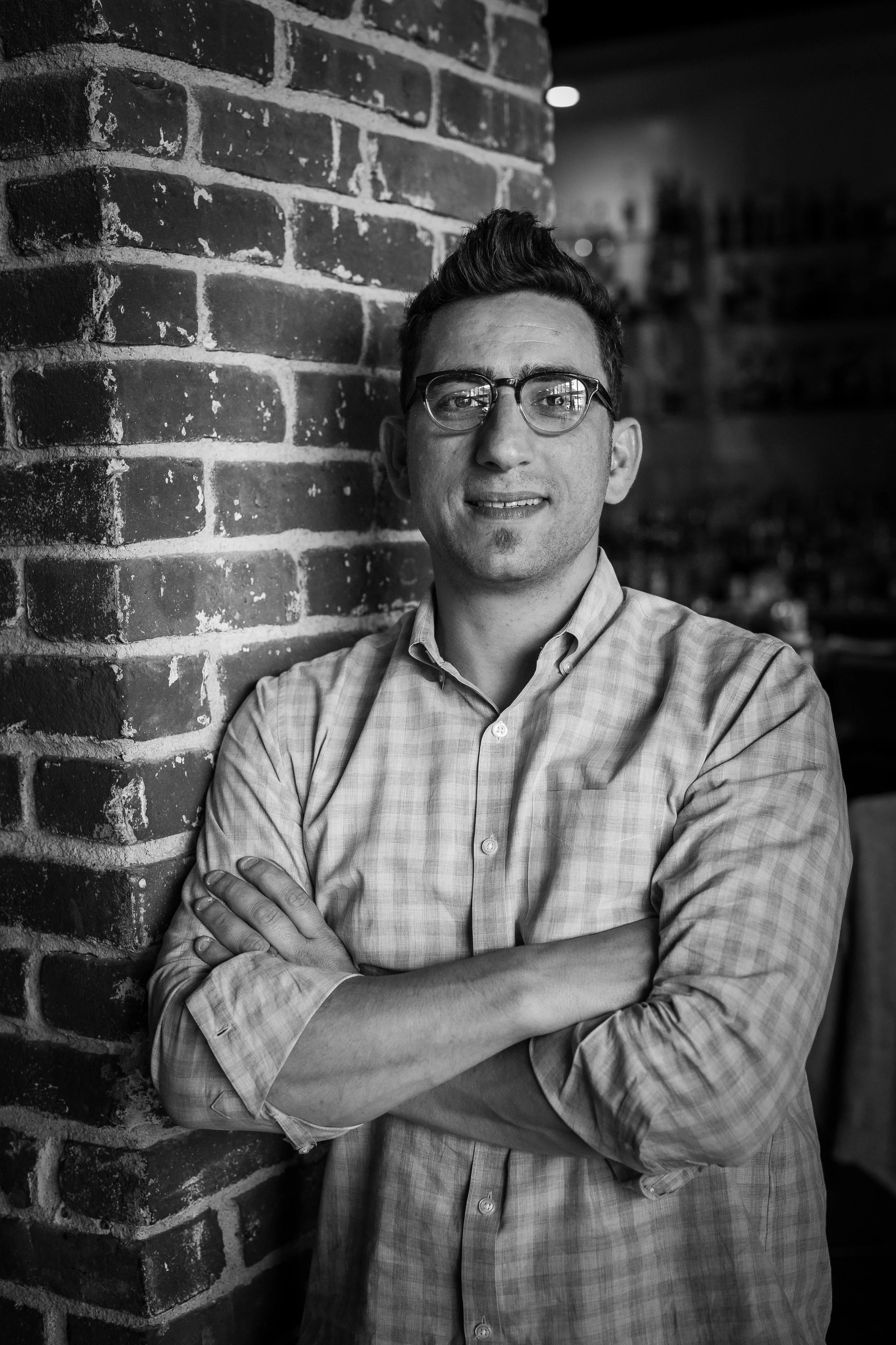 A man with short dark hair, glasses, and a slight beard, wearing a light checkered button-up shirt, standing with arms crossed, leaning against a brick wall in an indoor setting.