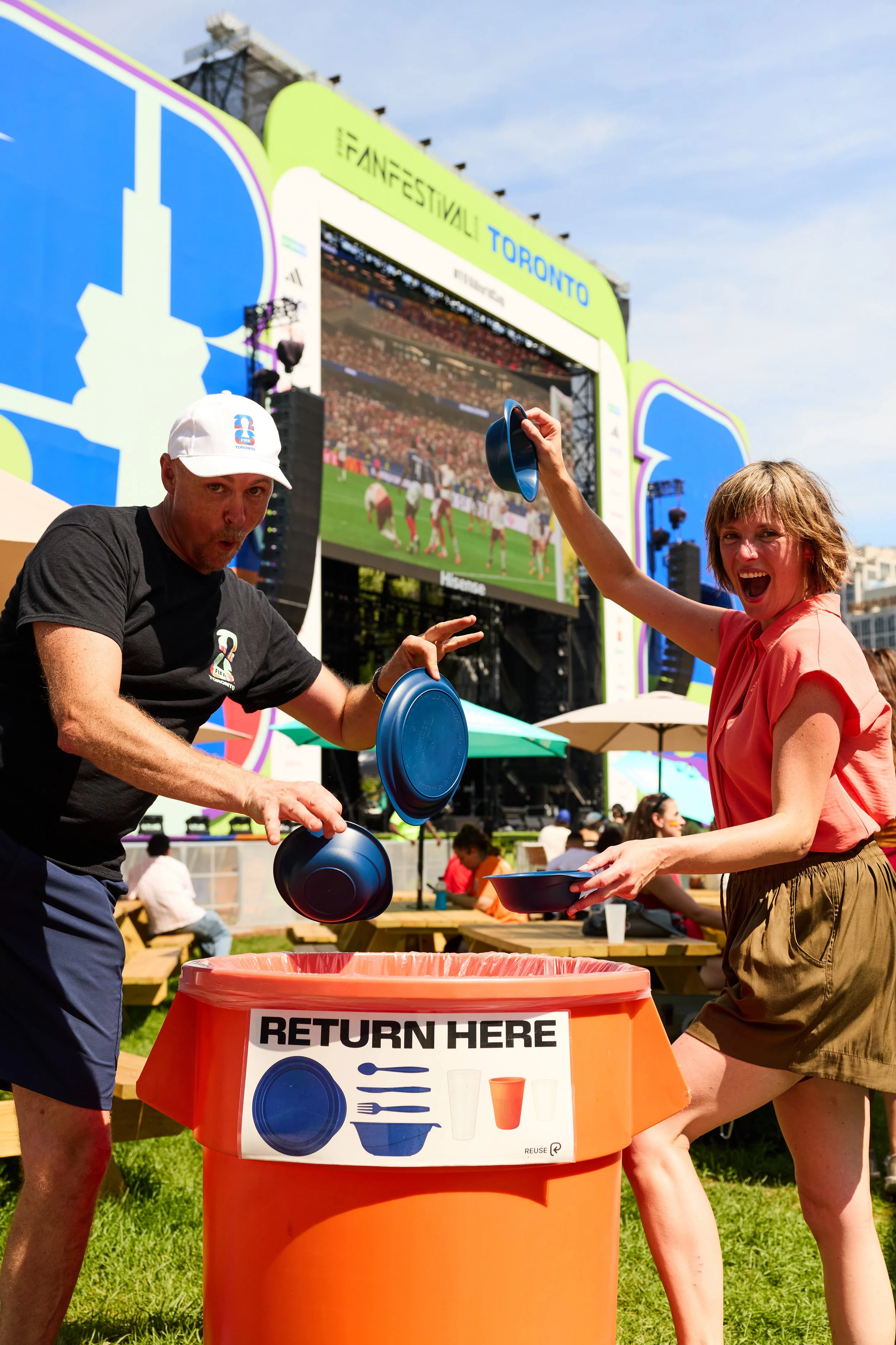 two people posing while putting reusable containers into a return bin at a FIFA fan fest