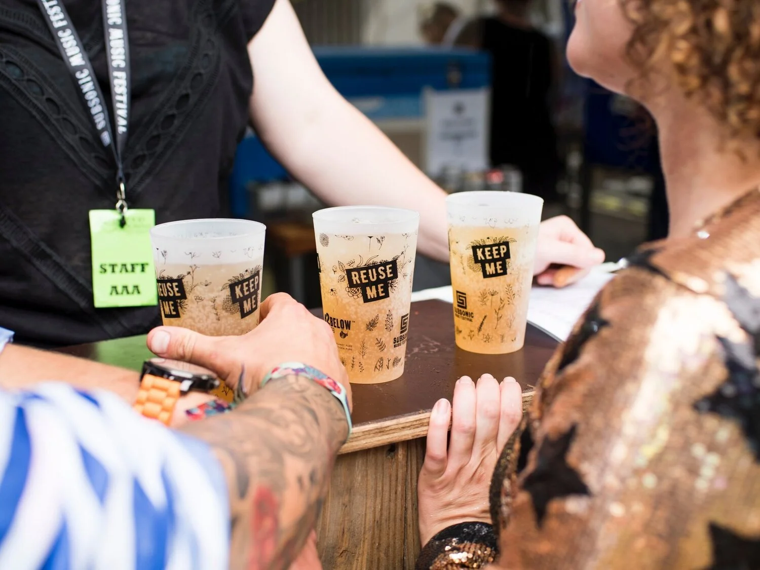Two people are holding reusable cups of beer at an event, with a staff member wearing a black shirt and a green staff badge in the background.