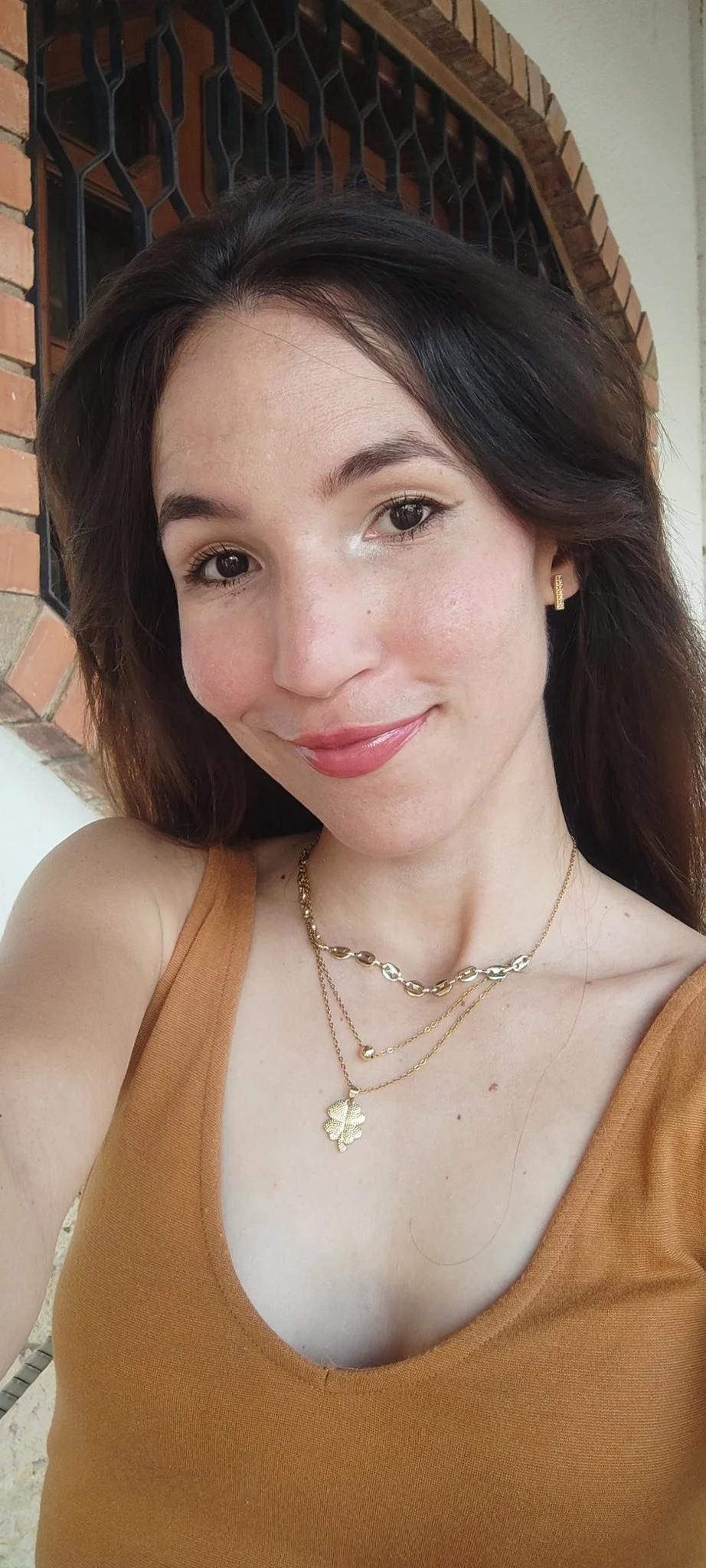 A woman with brown hair, wearing a caramel-colored tank top, gold jewelry including layered necklaces, hoop earrings, and a leaf pendant, smiling in front of a brick and iron window