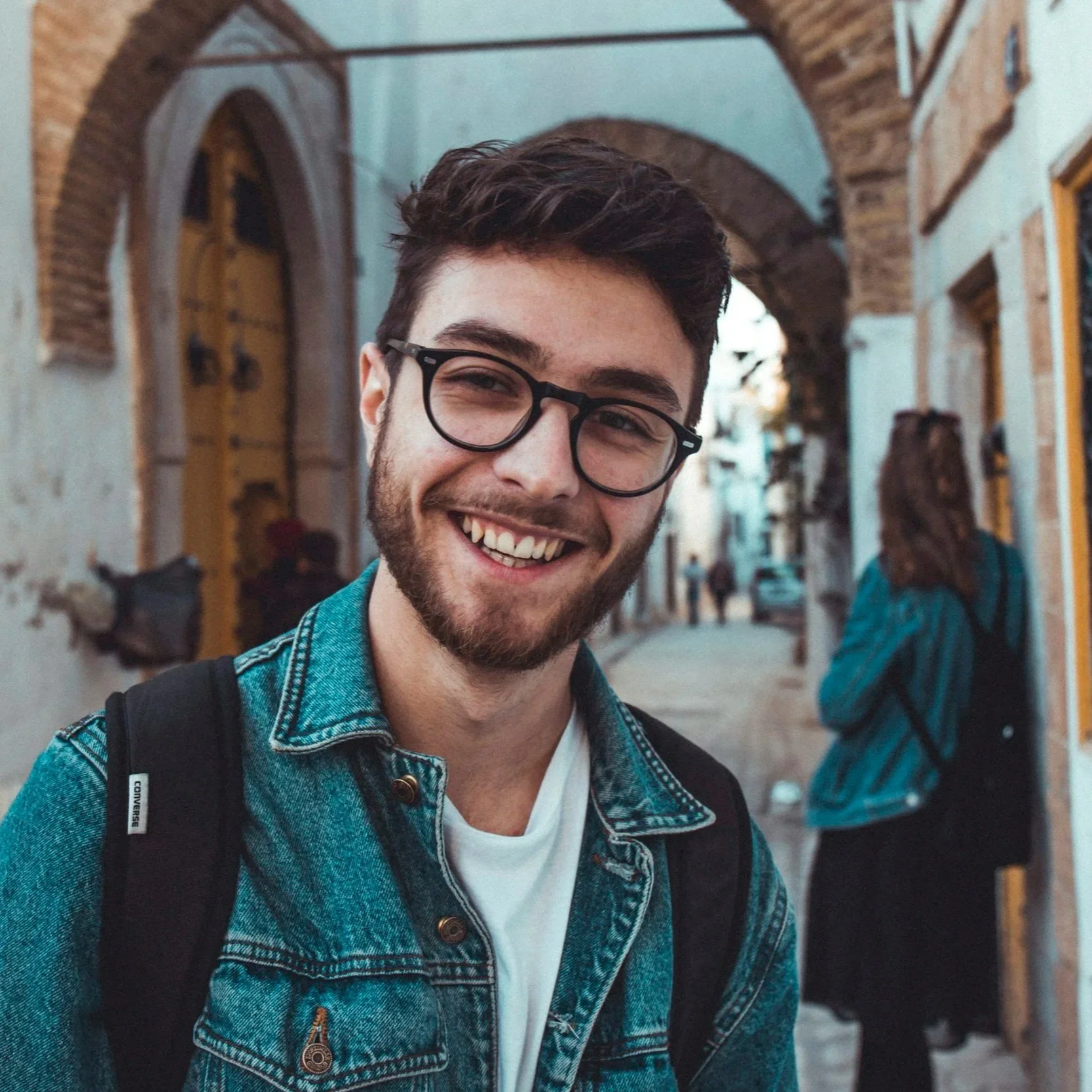 A young man with dark hair, glasses, and facial hair smiling at the camera, wearing a denim jacket and a white shirt, in a street with brick arches and an arched yellow door in the background.