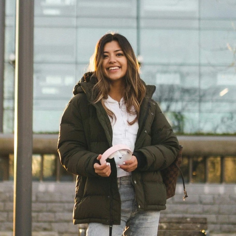 A young woman with long dark hair, smiling, standing outdoors with glass buildings behind her. She is wearing a black puffer jacket, white shirt, and light jeans, holding a pink object in her hand.