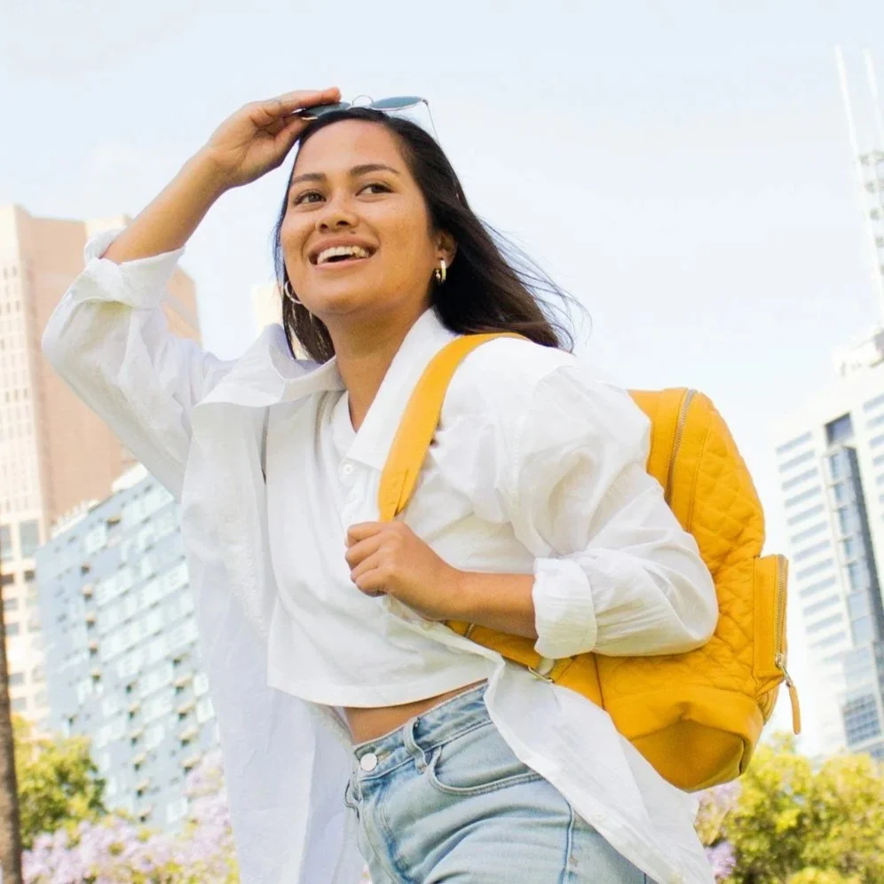 A young woman with long dark hair, smiling, wearing a white shirt and light blue jeans, holding a yellow backpack, with a cityscape in the background.