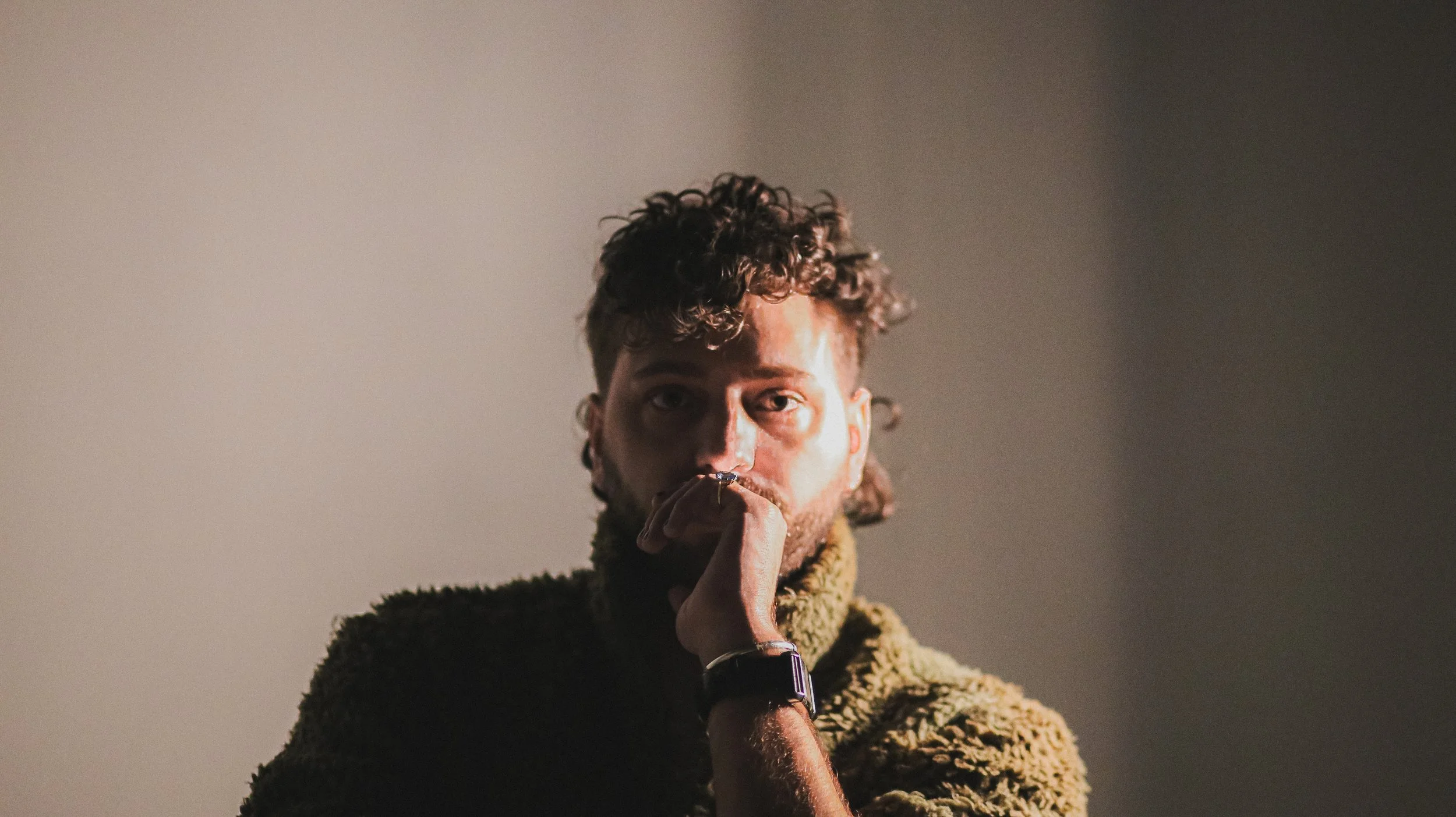 a photo of ryan, lit with a golden glow and a fuzzy green coat. He has a curly mullet, his great grandmother's aquamarine ring, a his late mother's bracelet, and is wearing a black watch.