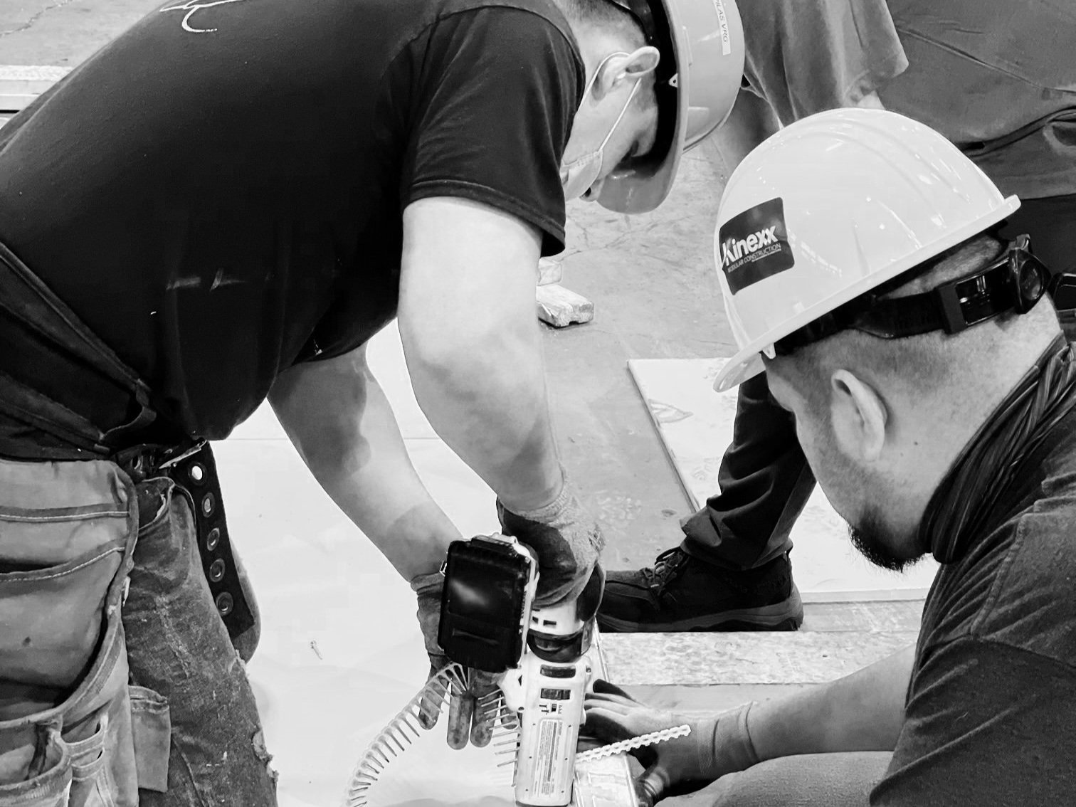Two construction workers wearing hard hats installing flooring using power tools.