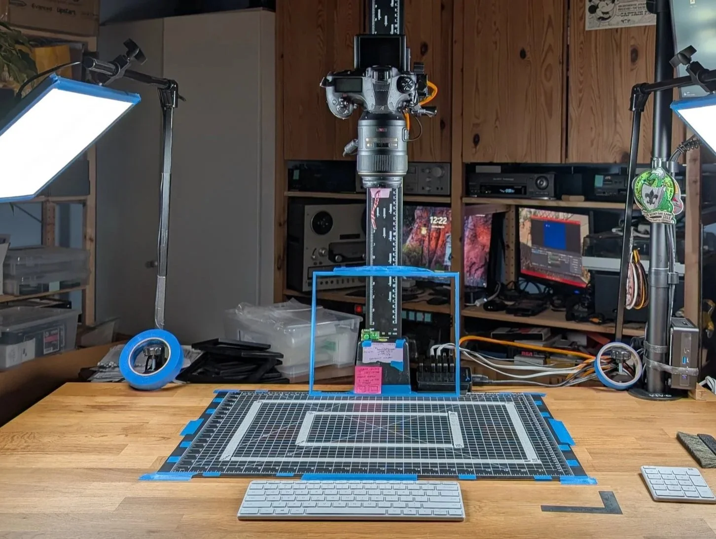 Professional overhead digitization station featuring a DSLR on a vertical column, aimed at a gridded cutting mat with taped registration marks. Dual LED panels provide cross-lighting, and surrounding tools include a keyboard, ruler, and monitor.