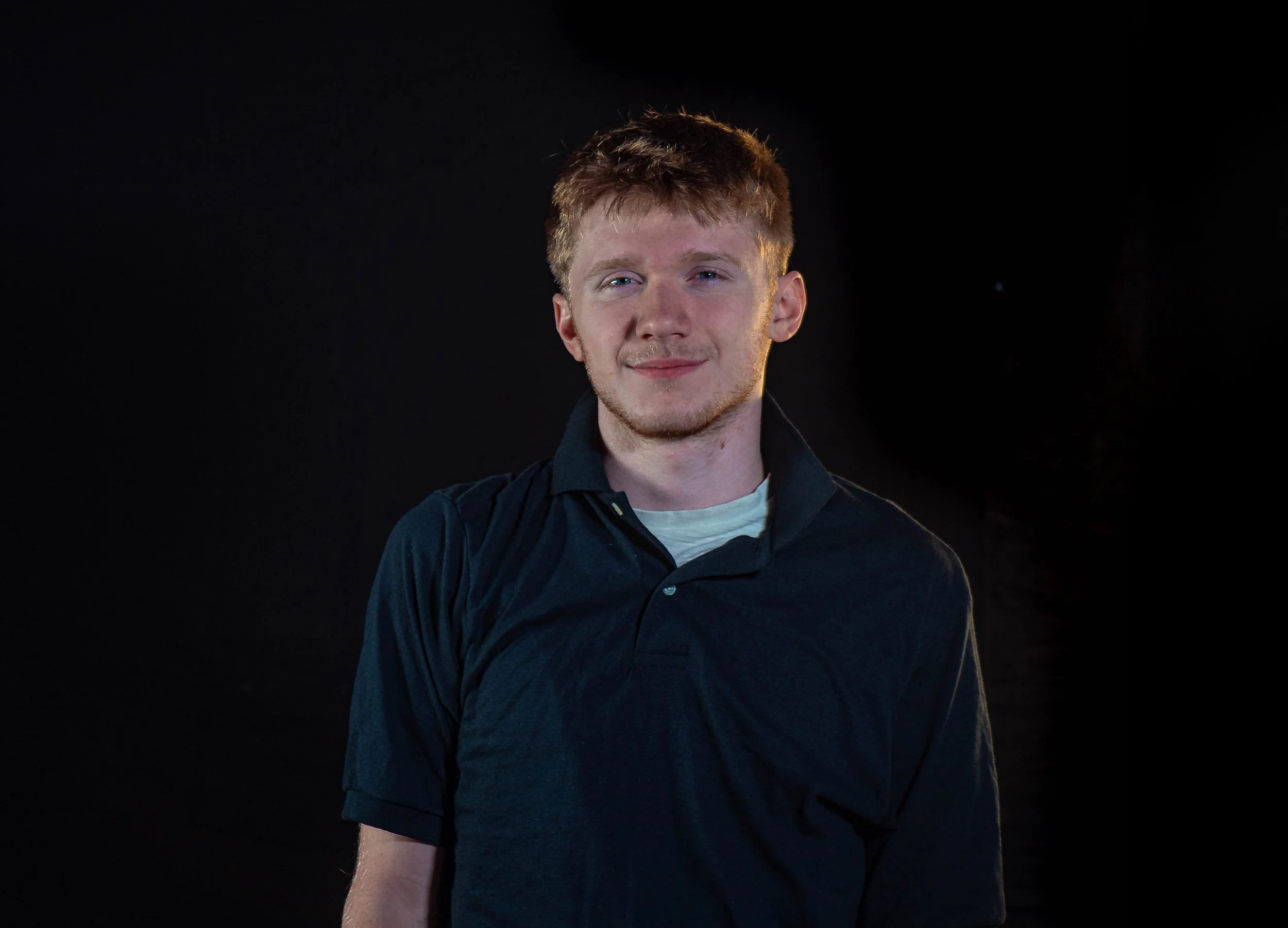 A young man with short light brown hair, light skin, and blue eyes stands against a black background. He is wearing a dark polo shirt over a white T-shirt and is smiling softly at the camera.