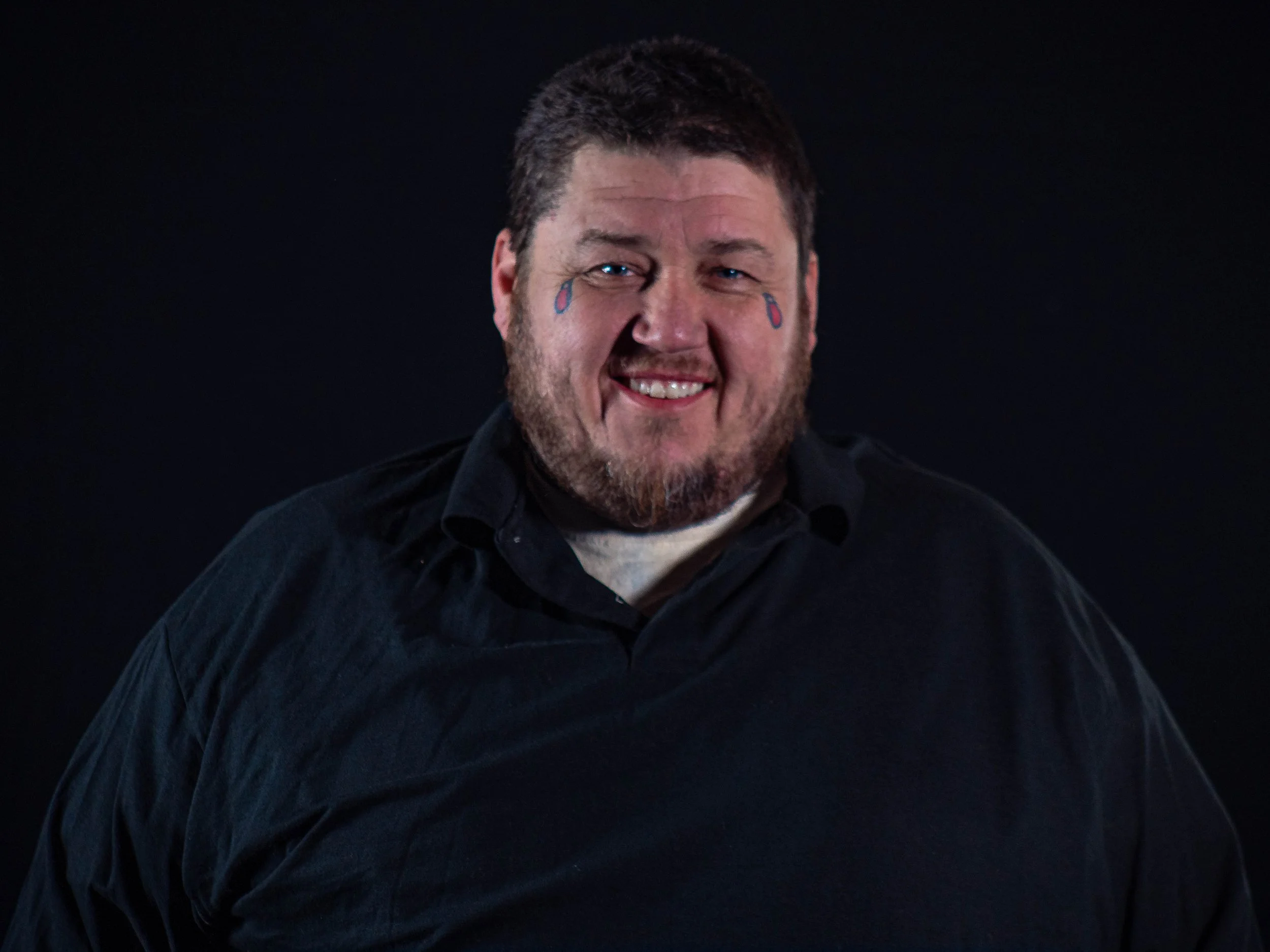 A smiling man with a beard and tattoos on his face, wearing a black shirt, against a dark background.