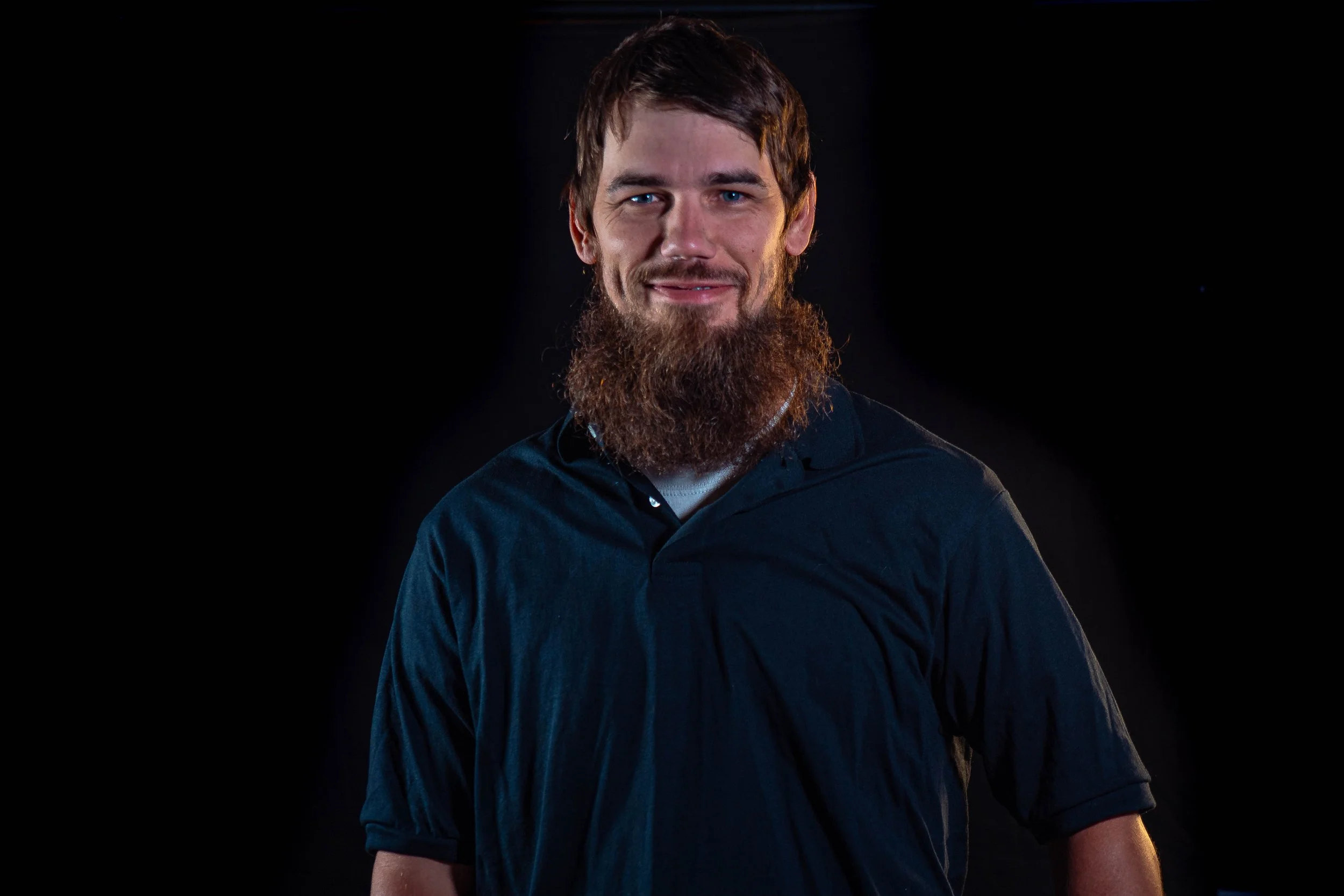 A smiling man with a beard and blue eyes wearing a black shirt against a dark background.