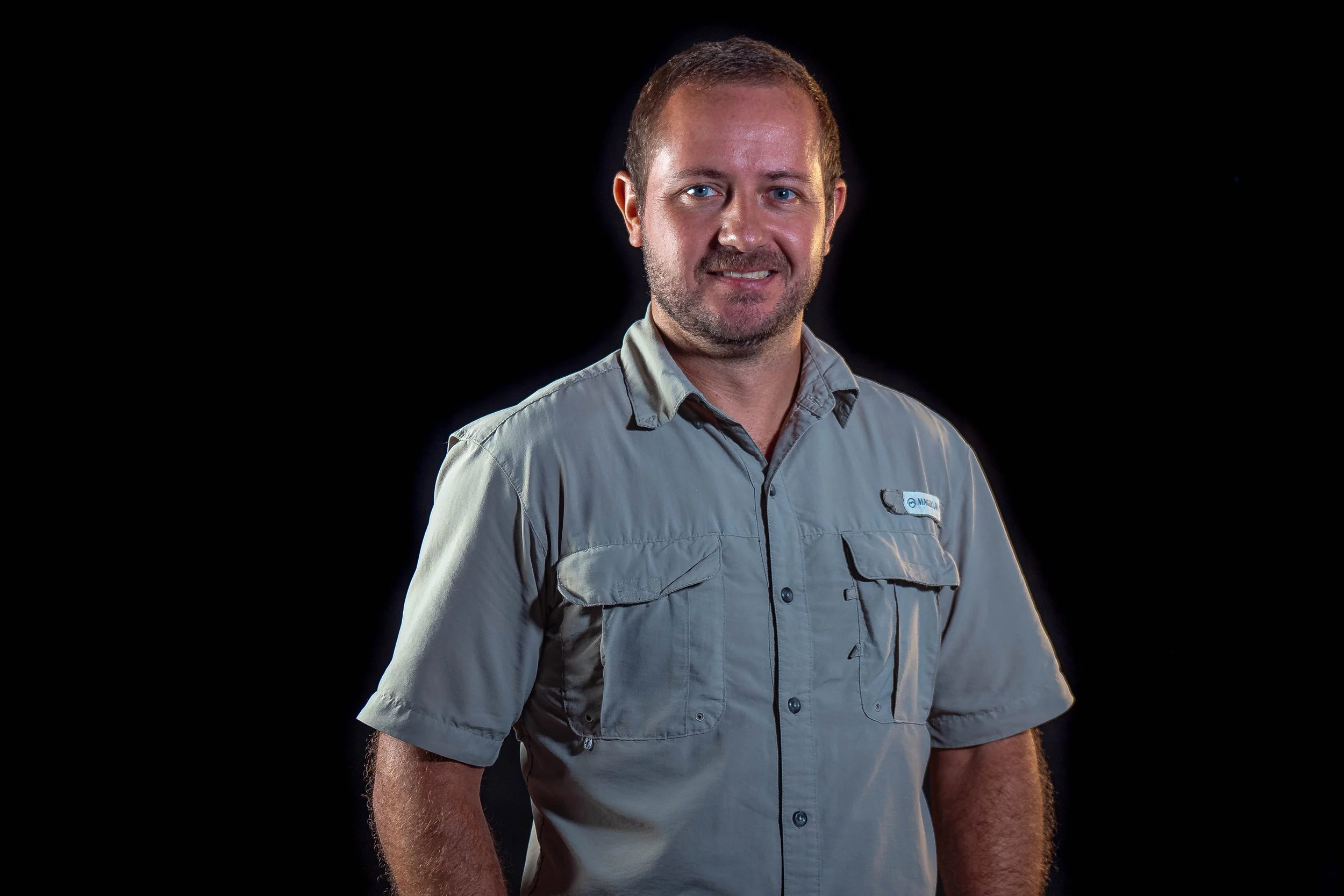 A man with short hair and a beard, wearing a light gray outdoor shirt with multiple pockets, smiling and standing against a black background.