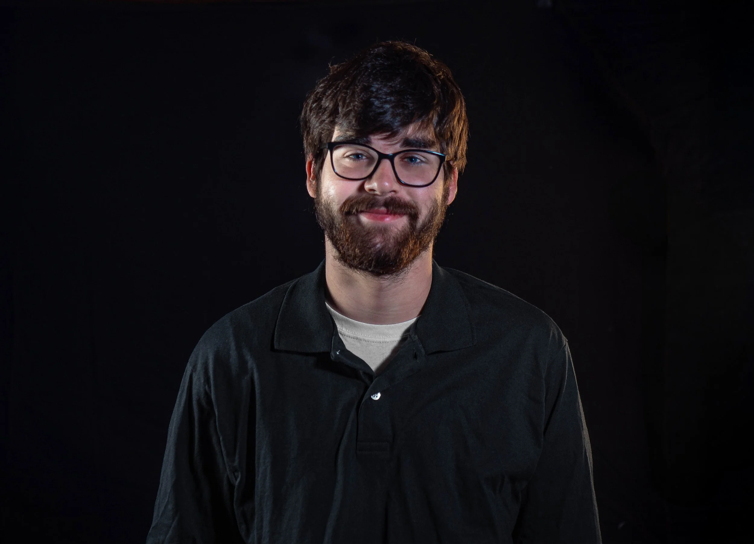 A young man with dark hair, glasses, and a beard, wearing a black jacket over a light-colored shirt, standing against a dark background, smiling softly.
