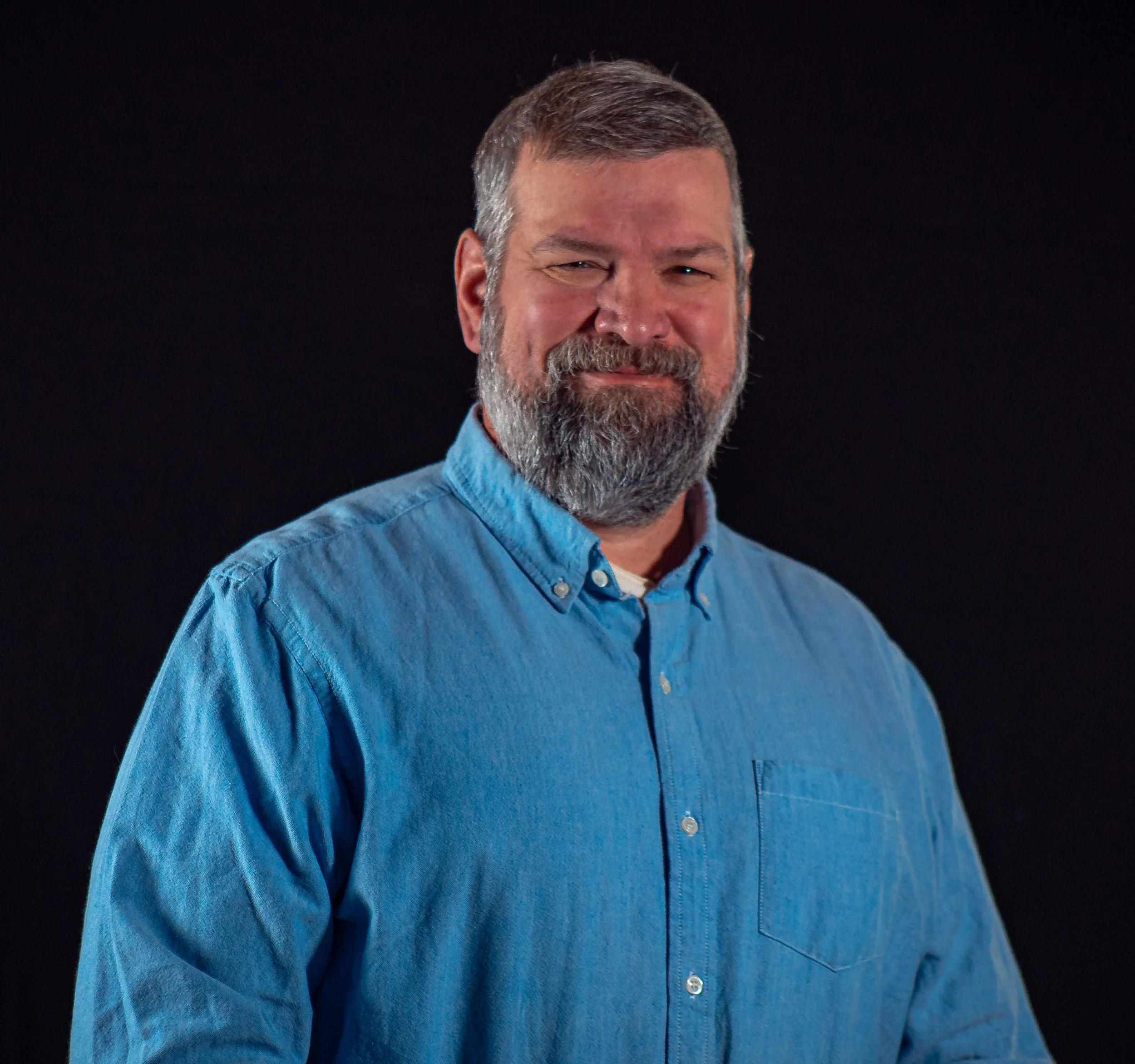 Portrait of a smiling man with a beard wearing a blue button-down shirt against a black background.