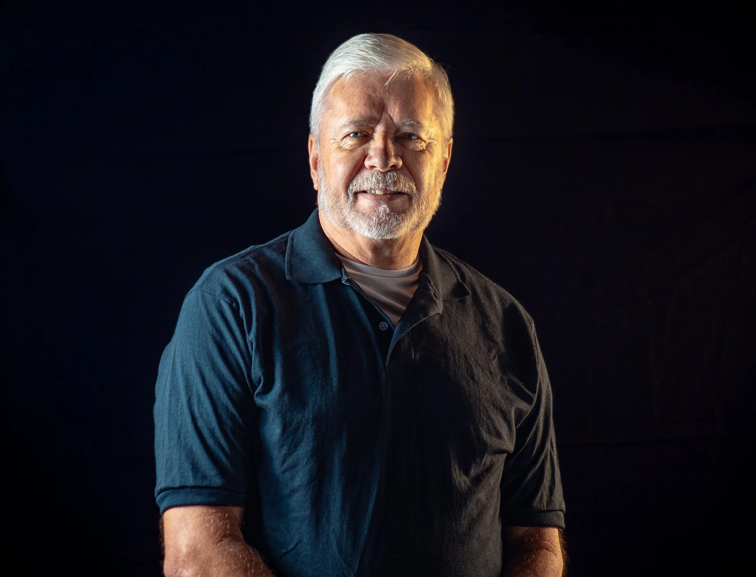 Portrait of an elderly man with white hair and beard, wearing a black polo shirt, smiling against a dark background.