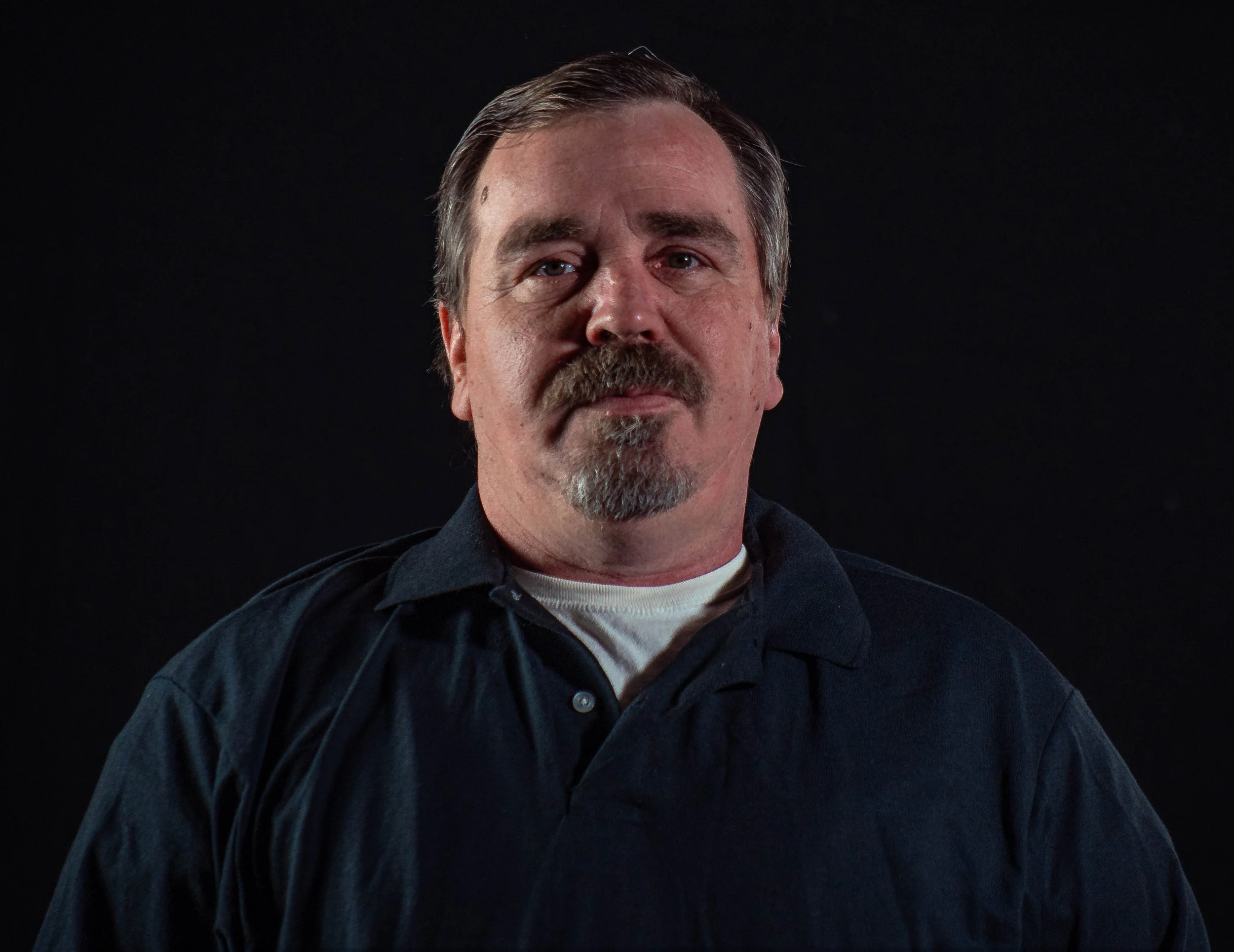A middle-aged man with gray hair and a goatee, wearing a black collared shirt over a white shirt, standing against a black background.