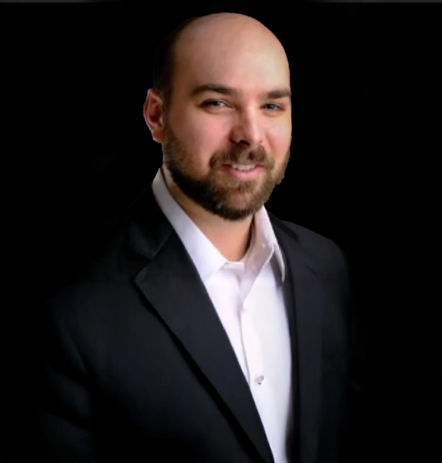 A man with a beard and bald head, wearing a black suit and white shirt, smiling against a black background.