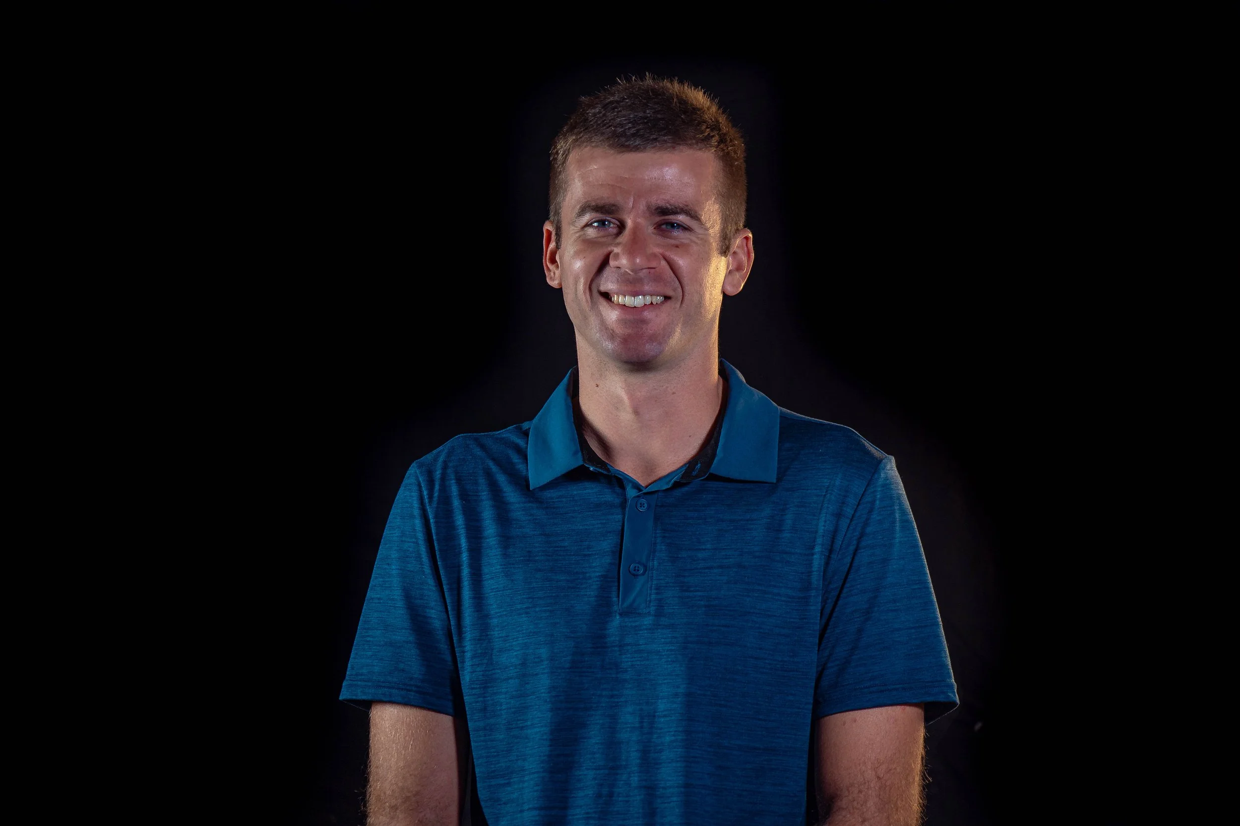 A man smiling in a dark studio, wearing a green polo shirt.