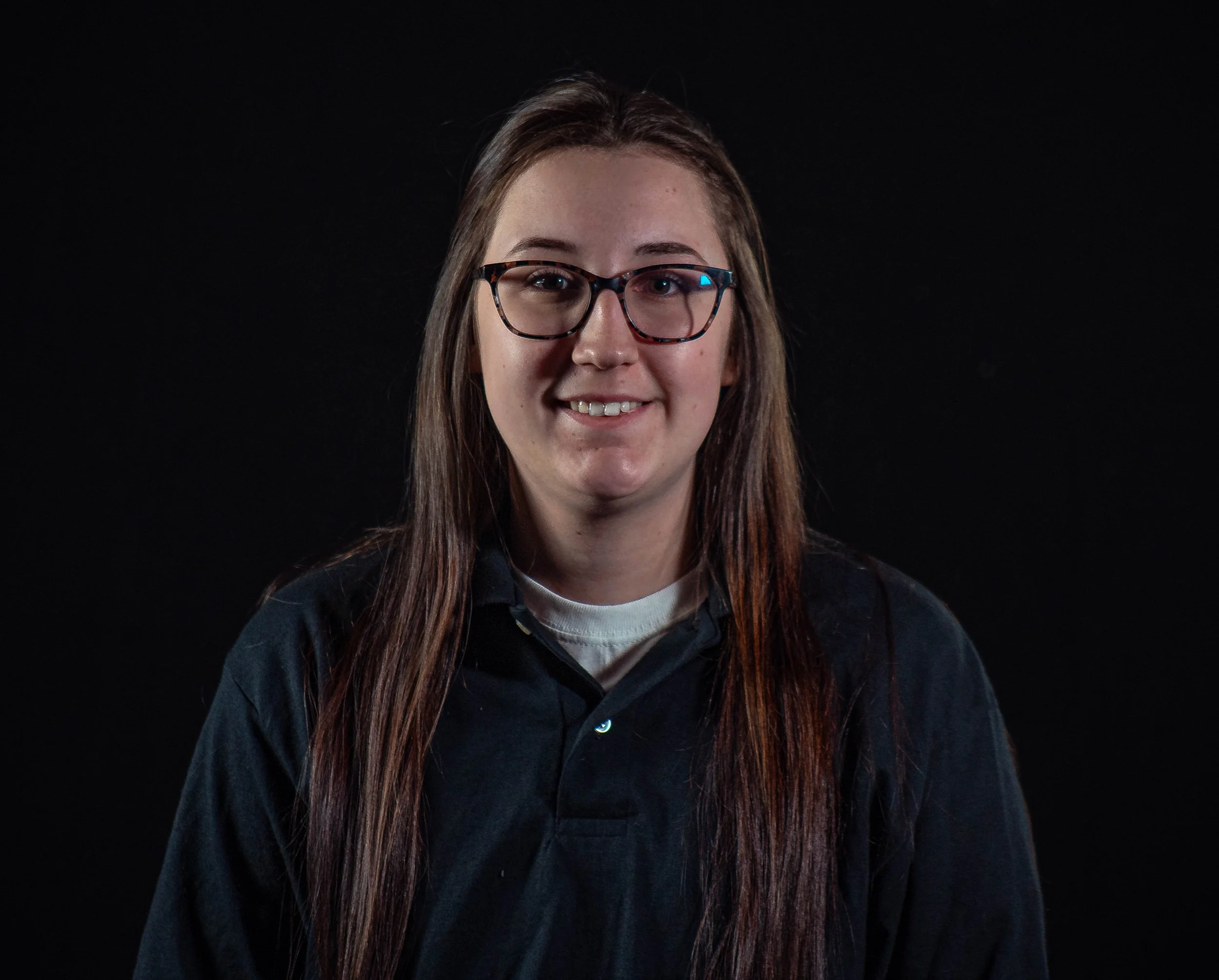 Portrait of a young woman with long brown hair, glasses, wearing a black shirt, smiling against a black background.