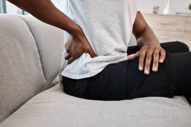 Person sitting on a gray couch, holding their lower back with one hand and resting the other hand on their thigh, indicating back pain or discomfort.