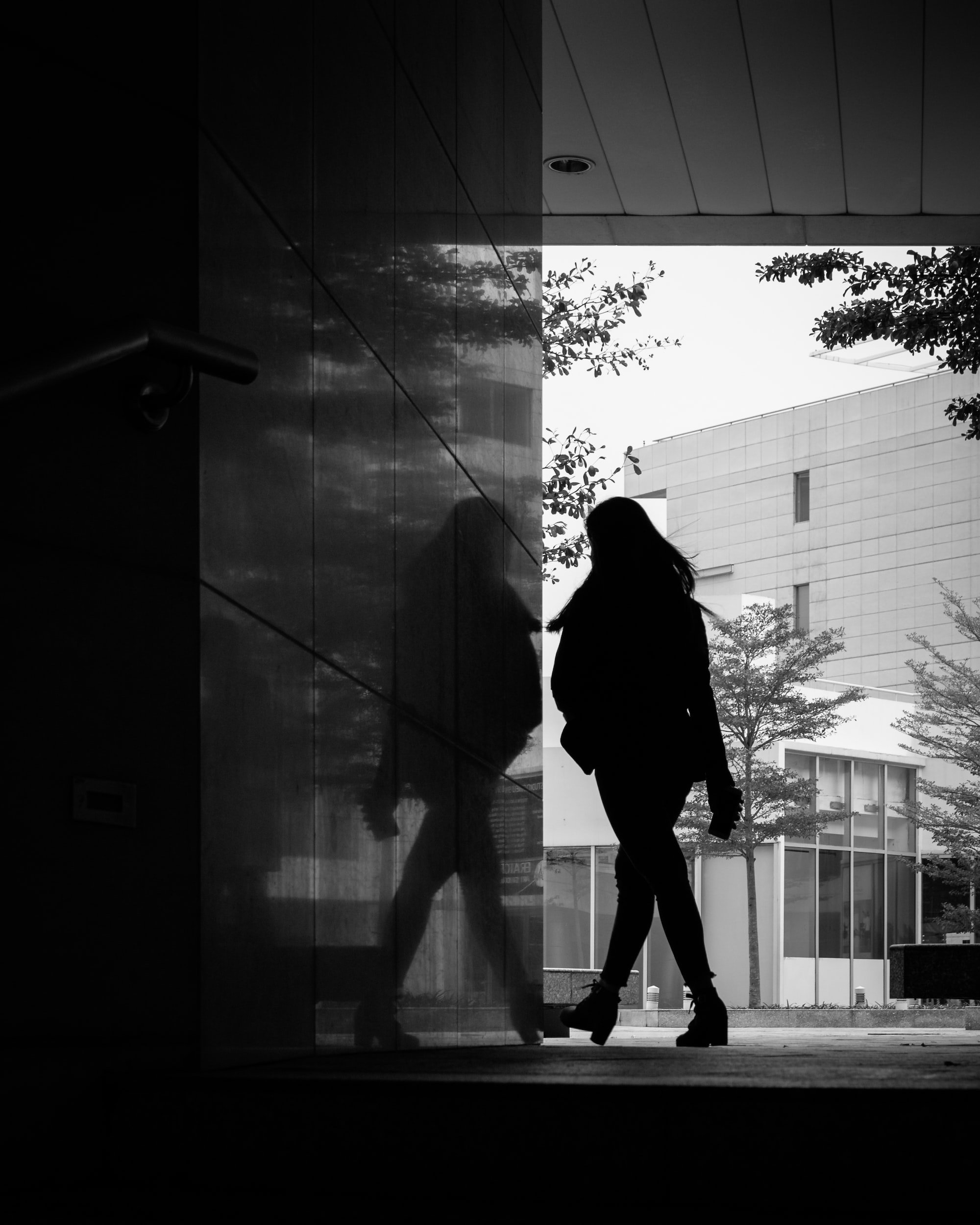 Silhouette of a woman walking outdoors near a modern building, with her reflection visible on a glass wall, in black and white.