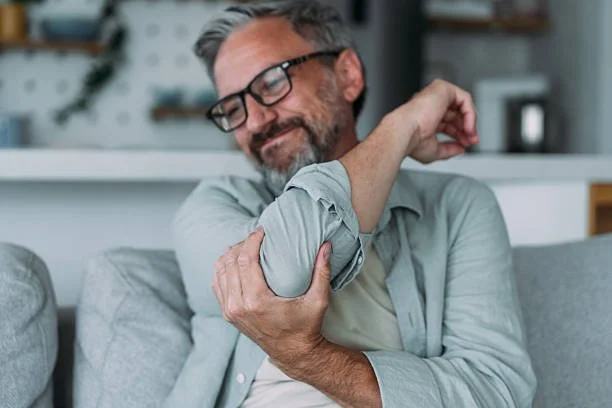 Man with glasses and gray hair sitting on a couch, smiling and rolling up his sleeve to show his arm.