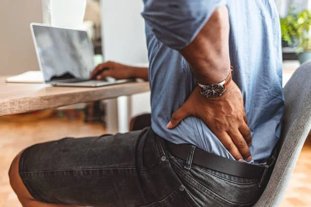A man sits at a desk with a laptop, holding his lower back in pain, indicating back discomfort.
