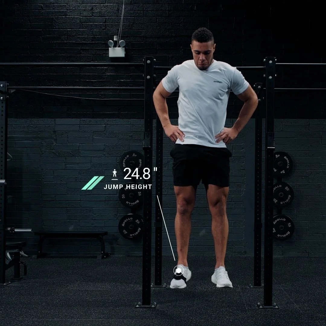 A man standing on a black gym floor between black weight racks, wearing a white t-shirt and black shorts, looking down with hands on hips. A digital overlay shows a jump height of 24.8 inches and a jump icon.