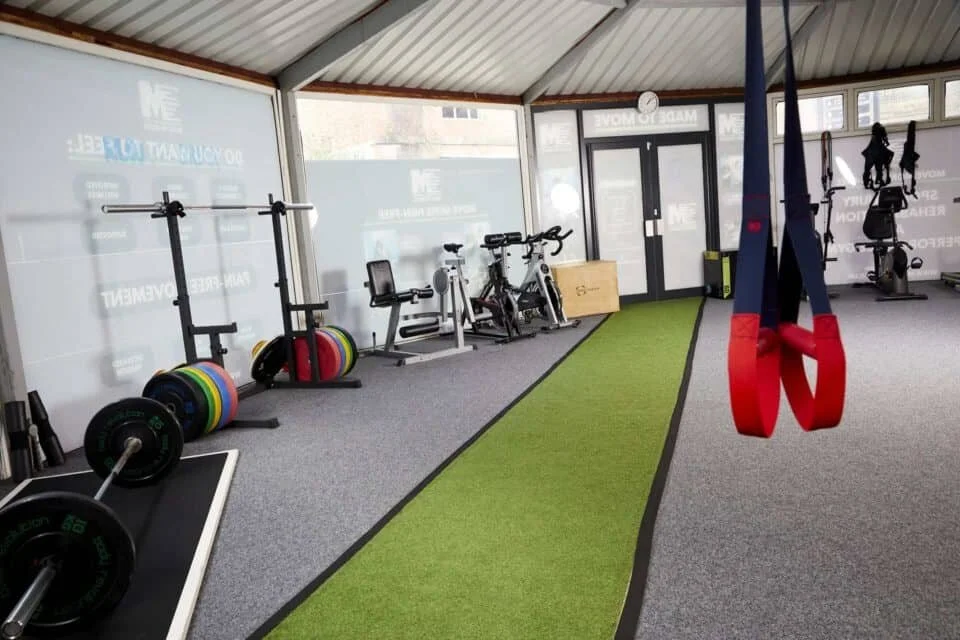 Indoor gym with weights, stationary bikes, and cardio equipment, featuring a green turf strip in the center, and red and blue suspension straps hanging from the ceiling.
