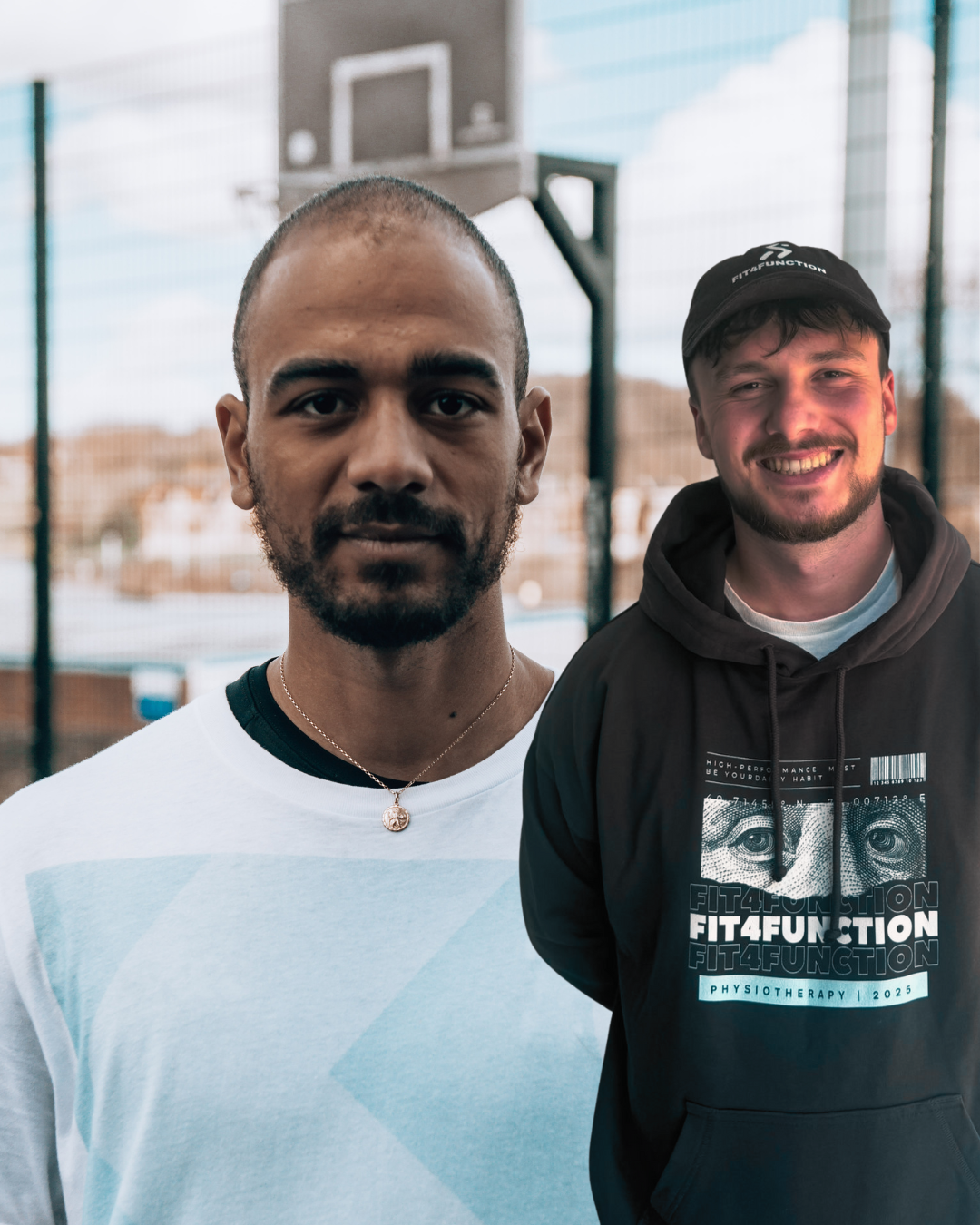 Two young men standing outside near a basketball court, smiling and facing the camera.