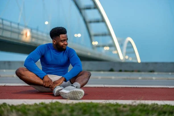 A man in a blue long-sleeve shirt and white shorts sitting on a red outdoor running track with a modern bridge in the background.