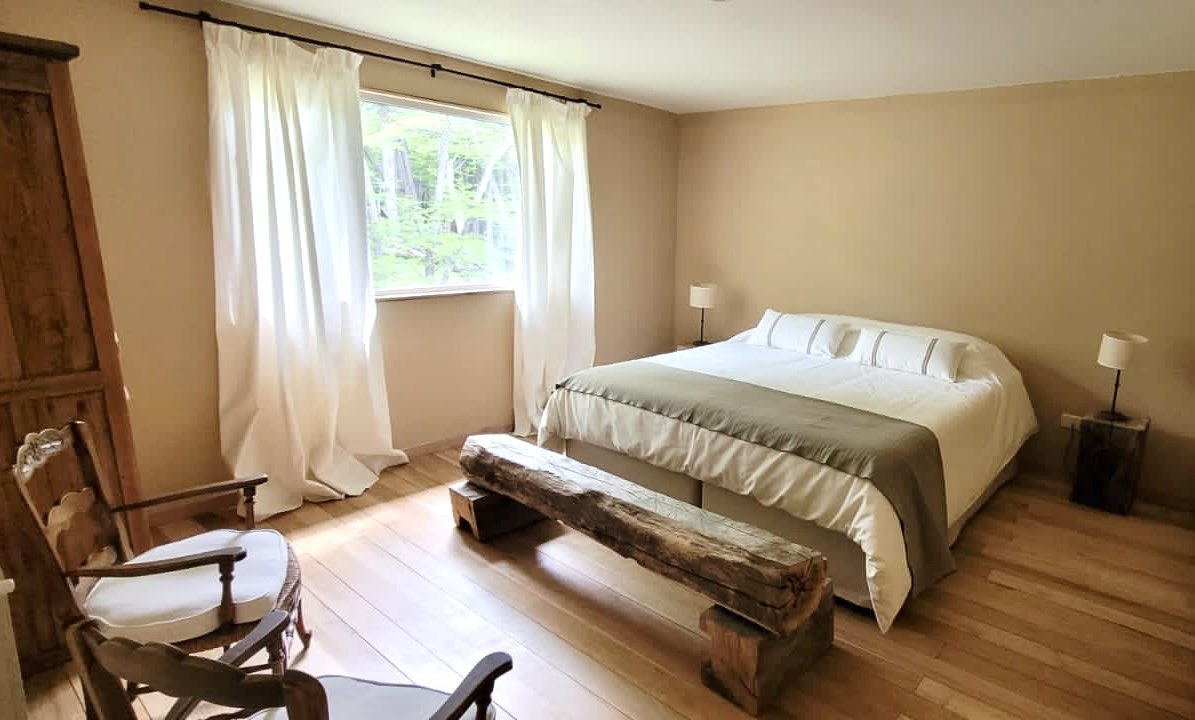 A bedroom with a bed, two side tables with lamps, a wooden armchair, and a rustic wooden bench, with a window and white curtains.