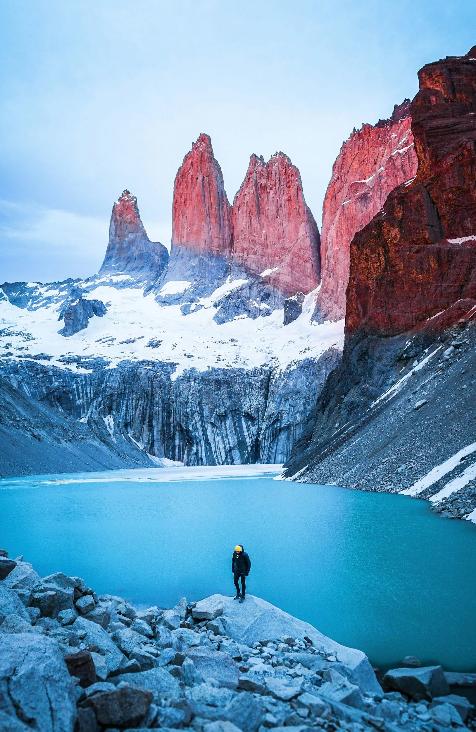 A person standing on rocks near a glacier-covered lake with towering red and dark mountain peaks in the background at sunset.