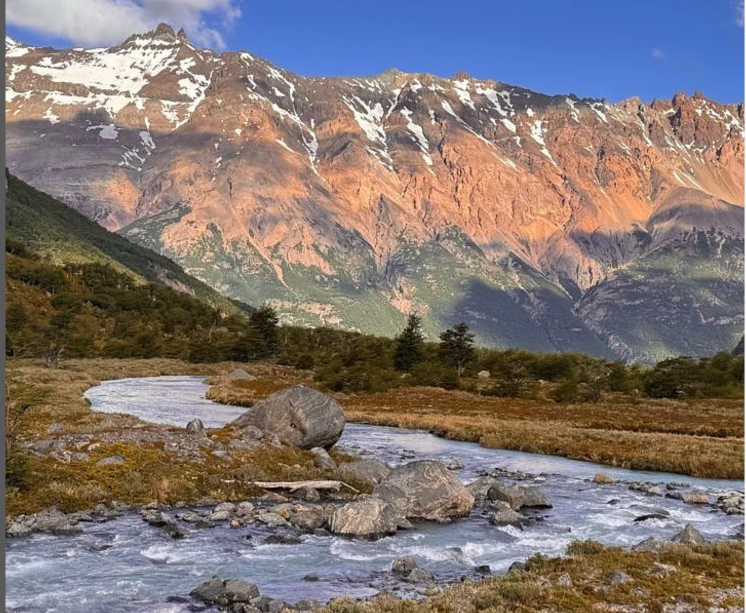 A mountain range with patches of snow, green forested slopes, rocky terrain, and a flowing river in the foreground under a blue sky.
