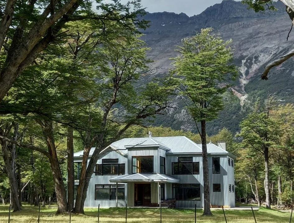 A two-story metal house with large windows, surrounded by trees and located at the base of mountains.