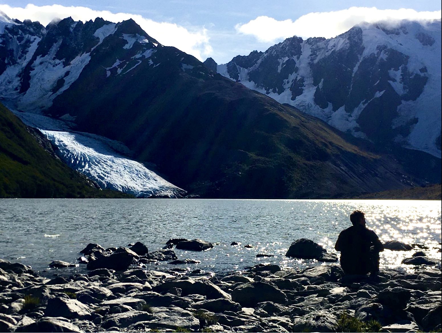 A person sitting on rocks by a lake, surrounded by snow-capped mountains and a glacier in the background, with partly cloudy sky.