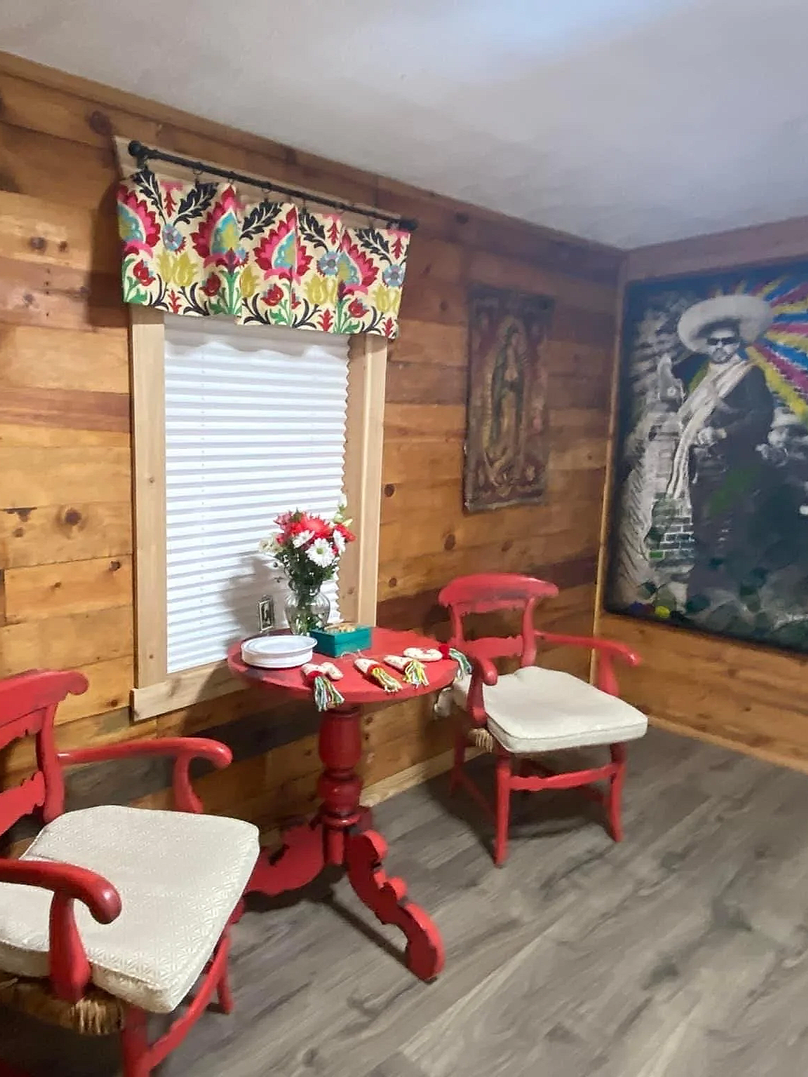 A cozy room with wood-paneled walls, a window with a floral valance and closed white blinds, two red wooden chairs with beige cushions, a small red table with a vase of flowers, decorative items, and colorful wall art.