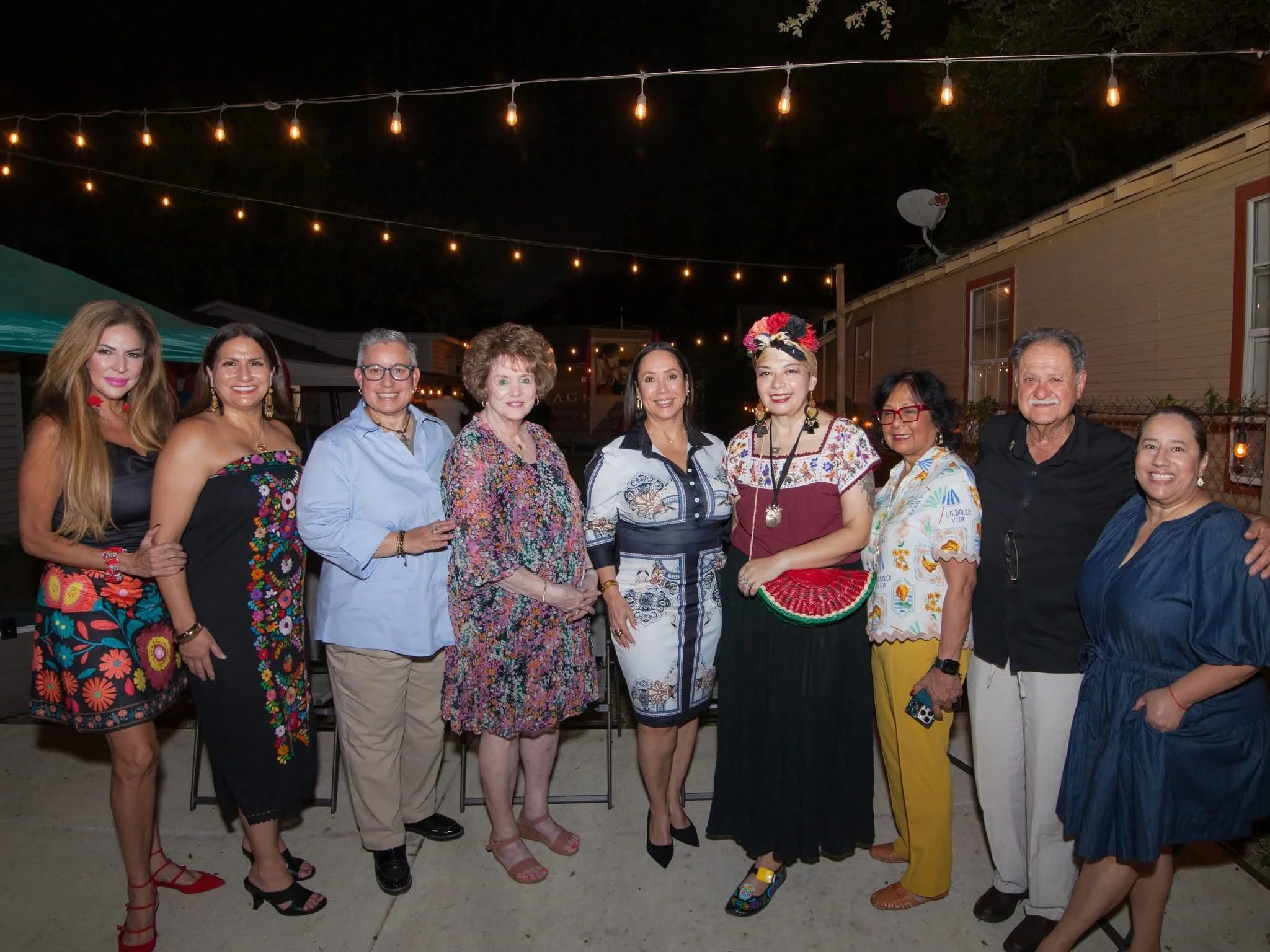 Group of nine people standing outdoors at night, smiling for a photo, with string lights hanging above.