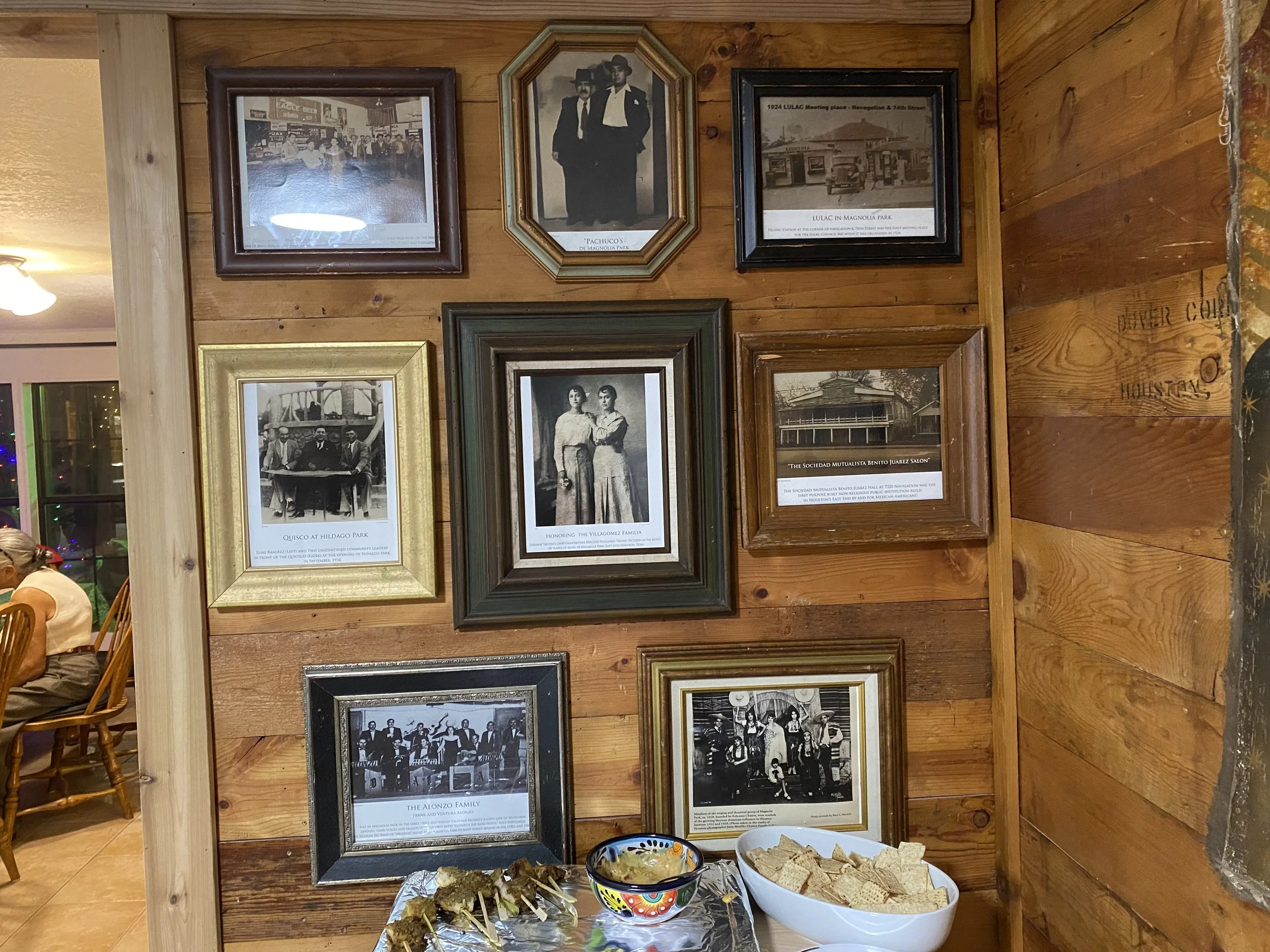 Close-up of a wooden wall decorated with framed black-and-white photographs and historical images, with a table of food including chips, nachos, and dips in the foreground.