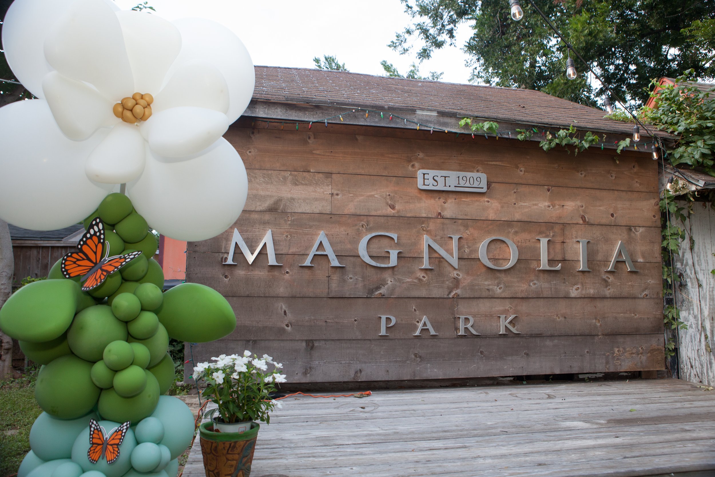 Decorative balloon arrangement featuring white, green, and light blue balloons, with butterfly stickers, in front of a wooden park sign with the text 'Magnoila Park' and an 'EST. 1909' plaque, outdoors with trees and hanging string lights.